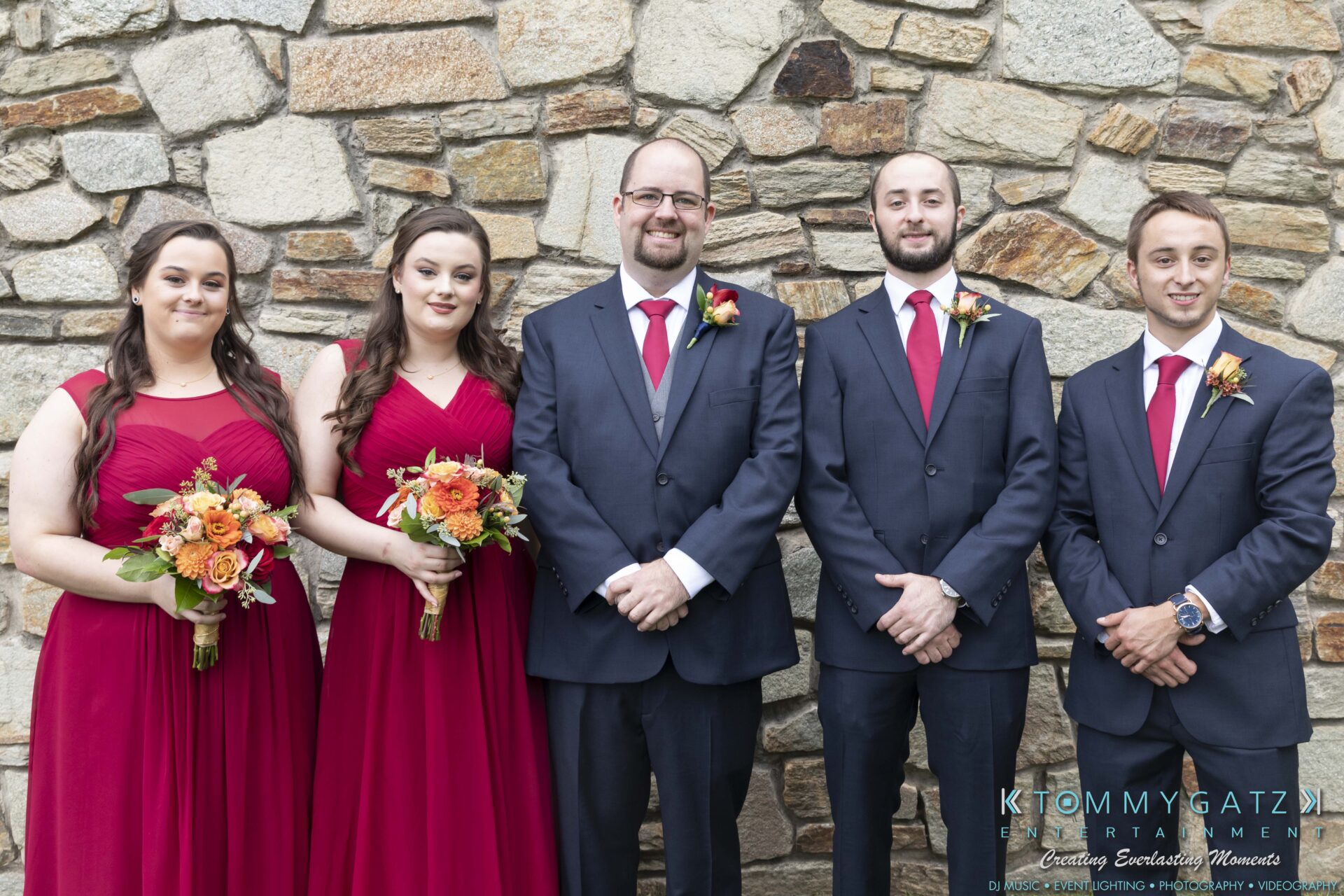 Five people in formal attire stand in front of a stone wall. Three women wear red dresses and hold bouquets. Two men wear dark suits with red ties.