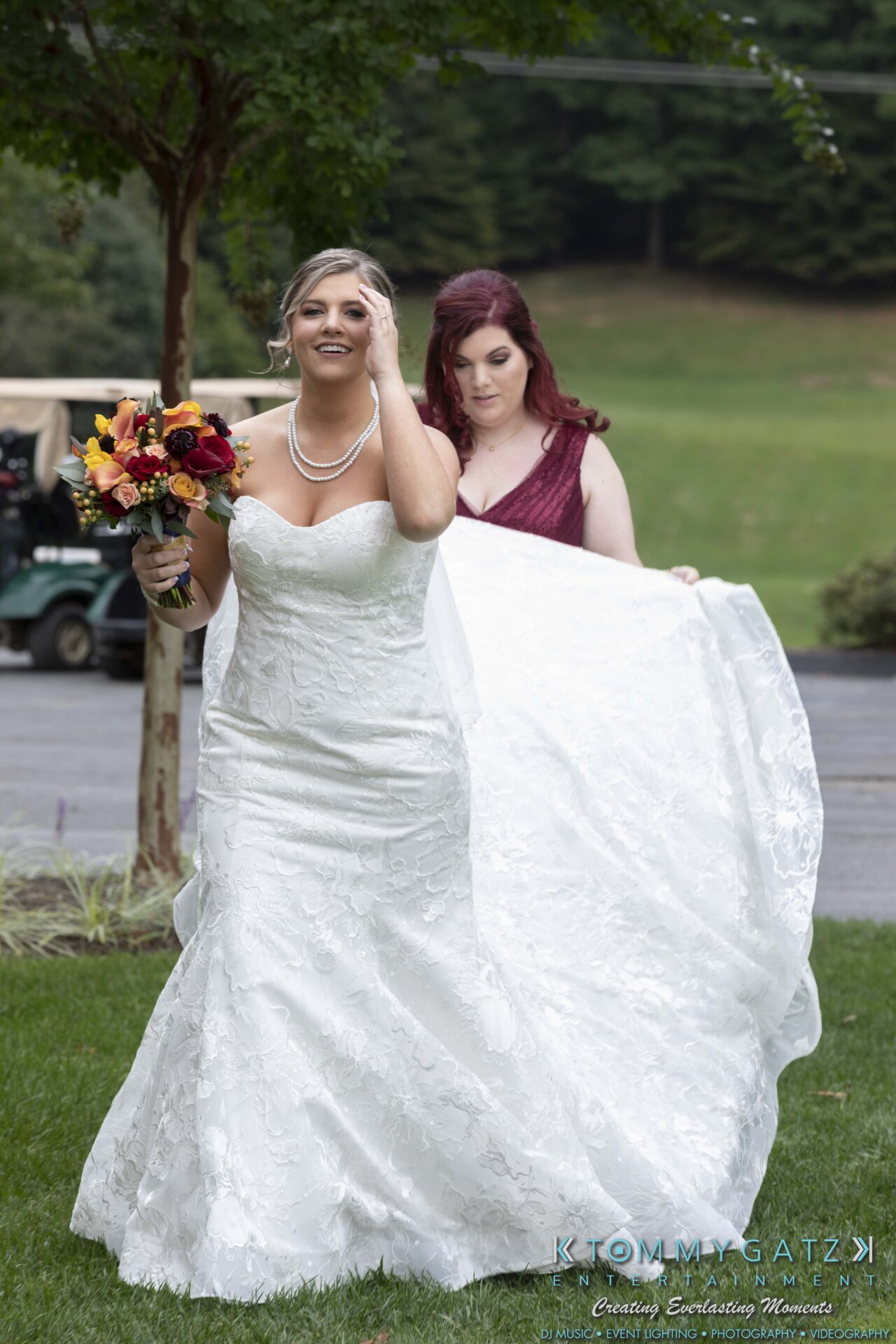 A bride holds a bouquet while a bridesmaid adjusts the train of her wedding dress outdoors.