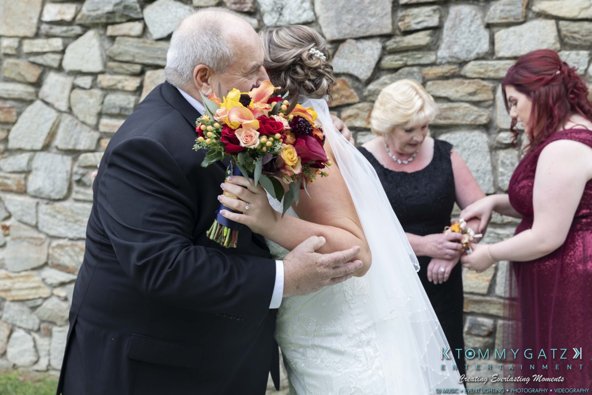 A bride in a white gown hugs a man in a suit holding a vibrant bouquet. Two women, one in black and one in red, stand nearby. A stone wall is in the background.