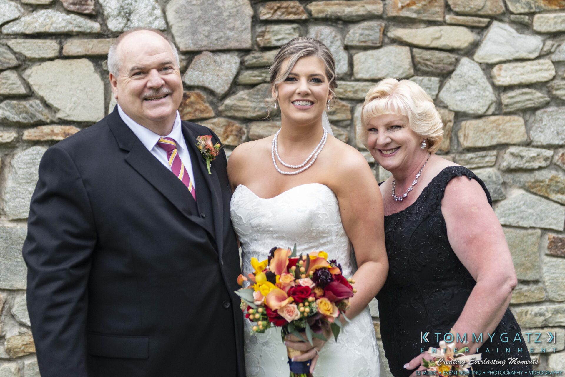 A bride in a white gown holds a bouquet, standing between two older adults in formal attire, in front of a stone wall.