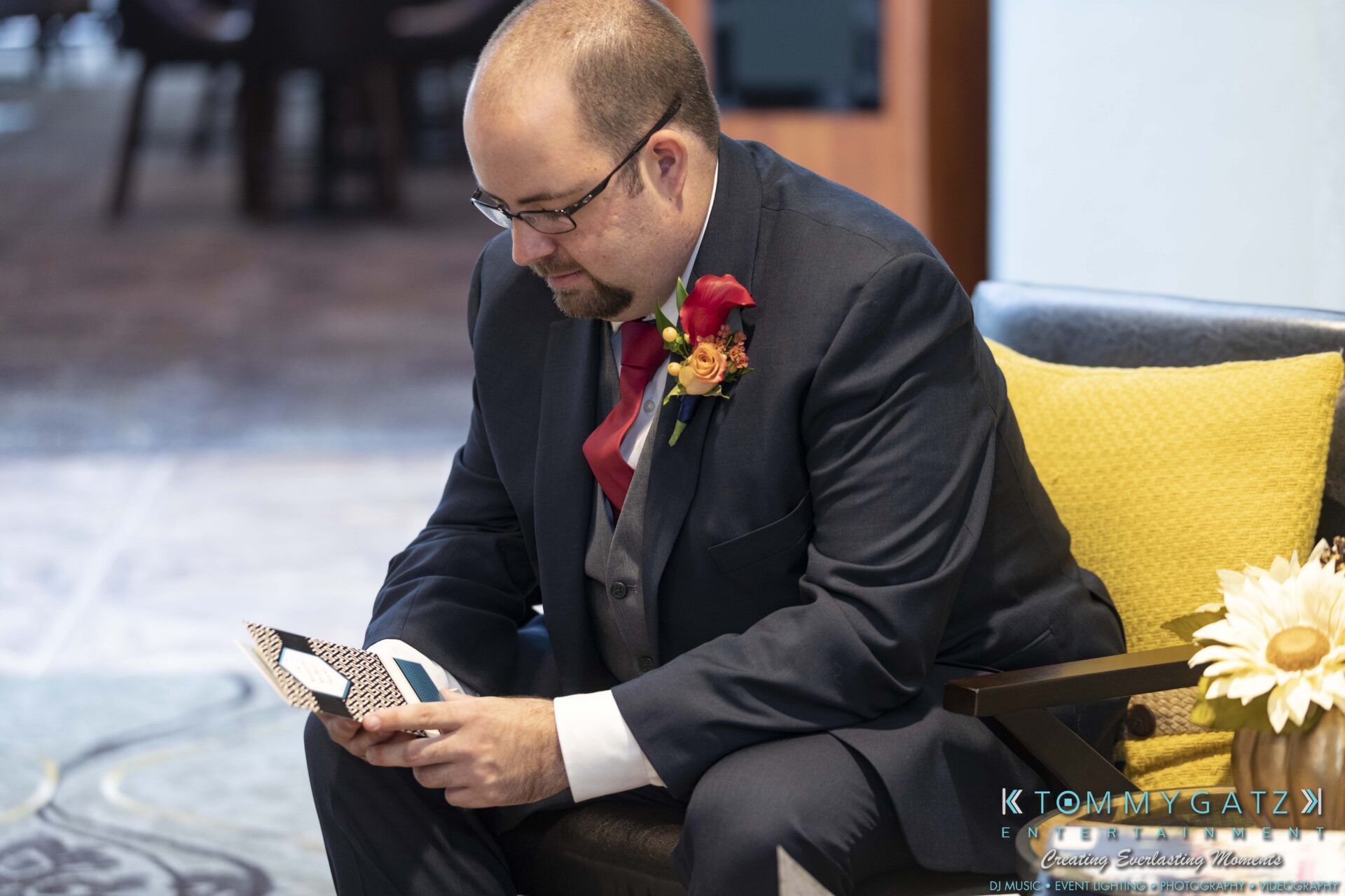 A man in a suit sits on a chair, holding a folded piece of paper, with flowers on his lapel and next to him.