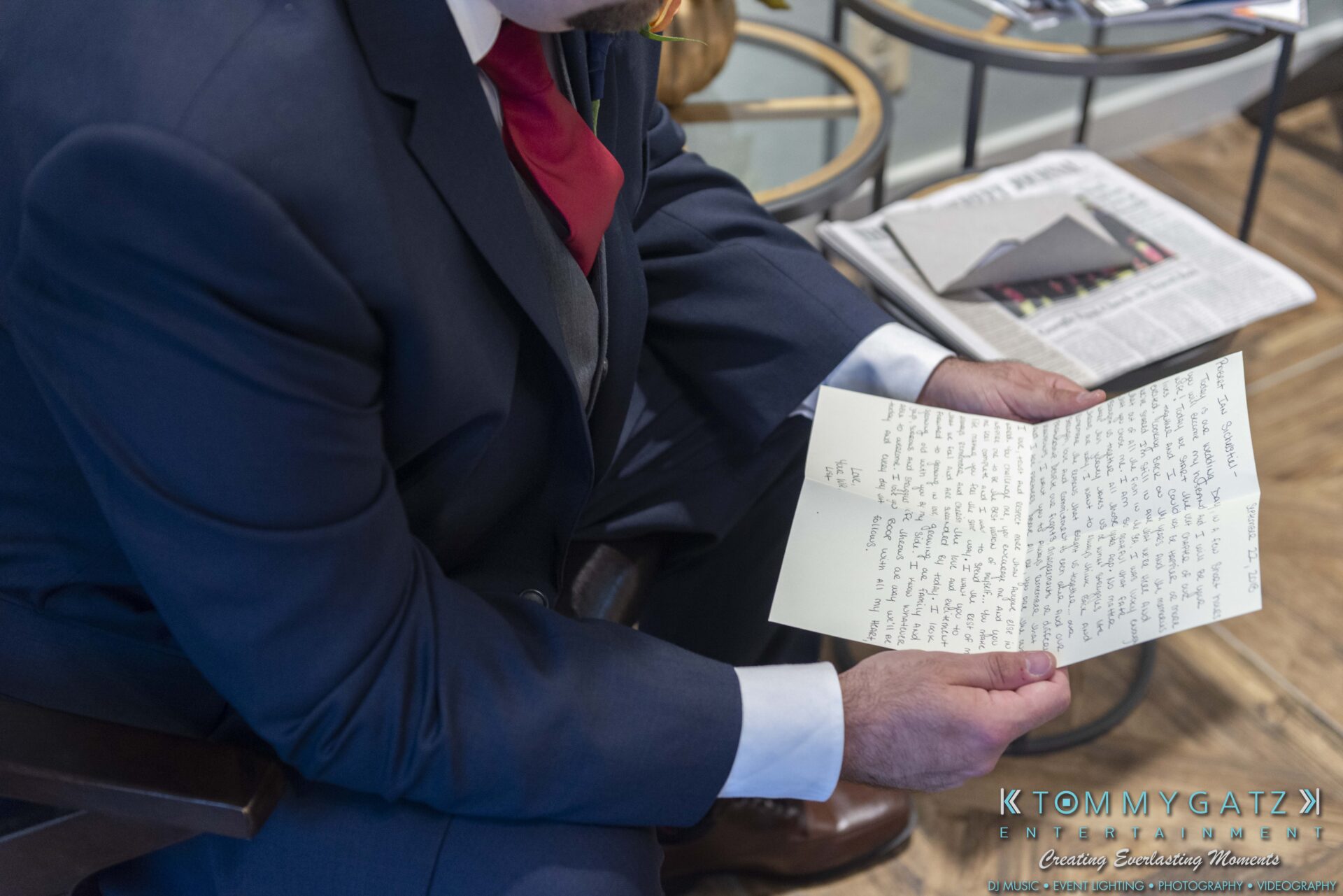 A person in a suit is sitting and reading a handwritten letter with a newspaper on the table nearby.