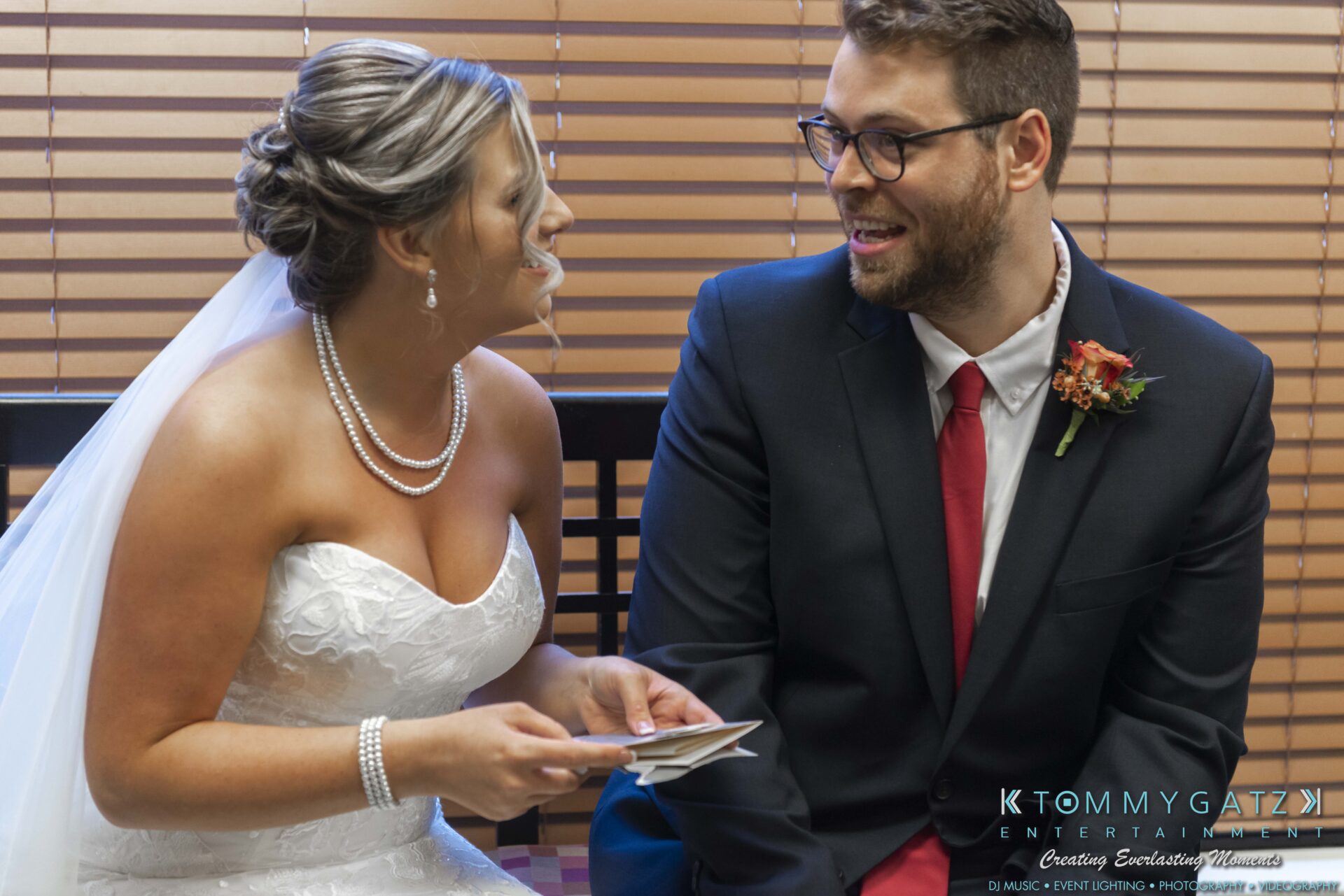 A bride in a white dress and veil sits beside a groom in a dark suit and red tie. They are smiling and holding notes, seated in front of window blinds.
