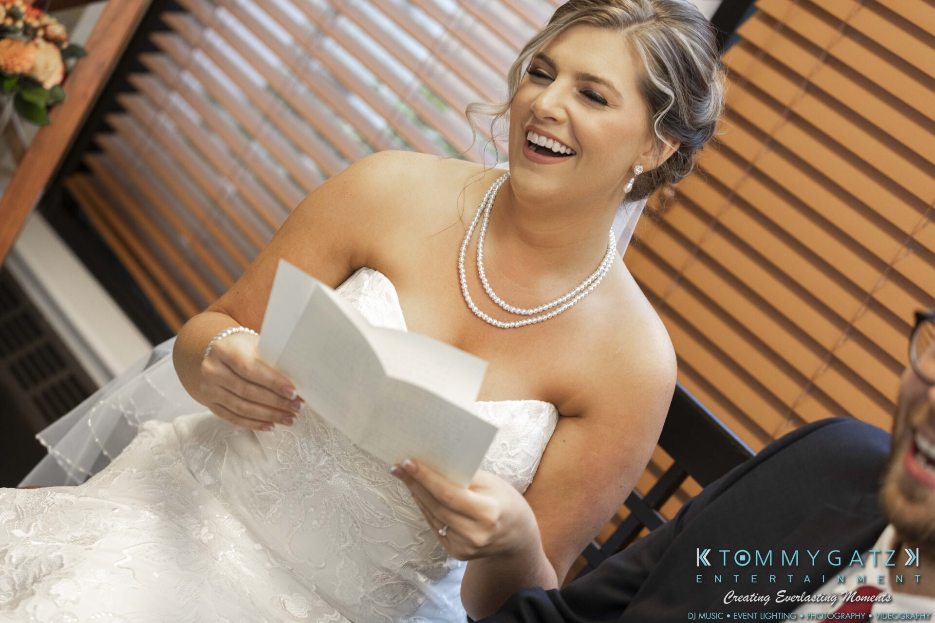 Bride smiling while reading a note, sitting next to a person. They're indoors, with blinds in the background and "KTOMMY GATZ ENTERTAINMENT" text on the image.