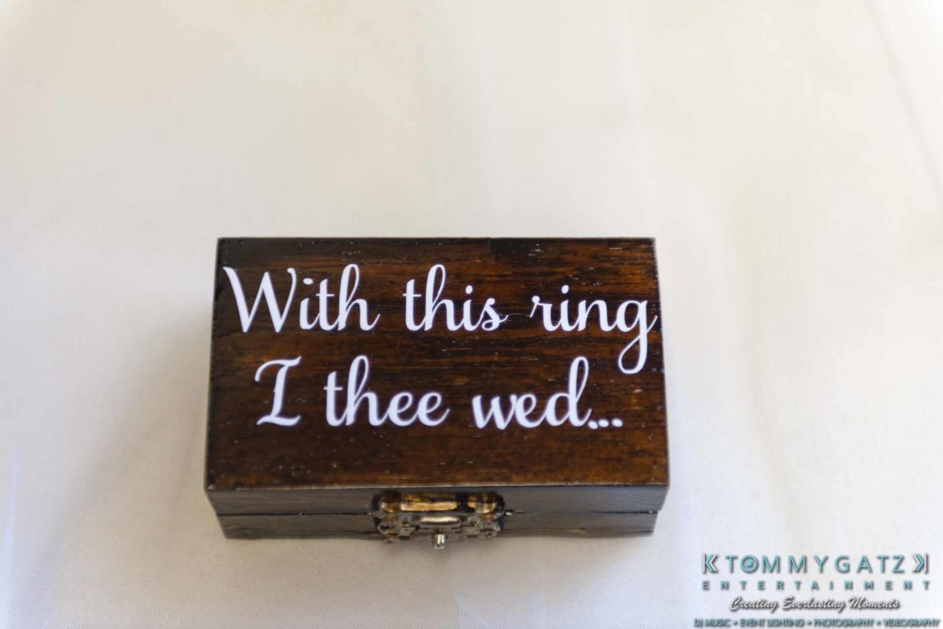A small wooden box with white text saying "With this ring, I thee wed" on the lid, placed on a white surface.