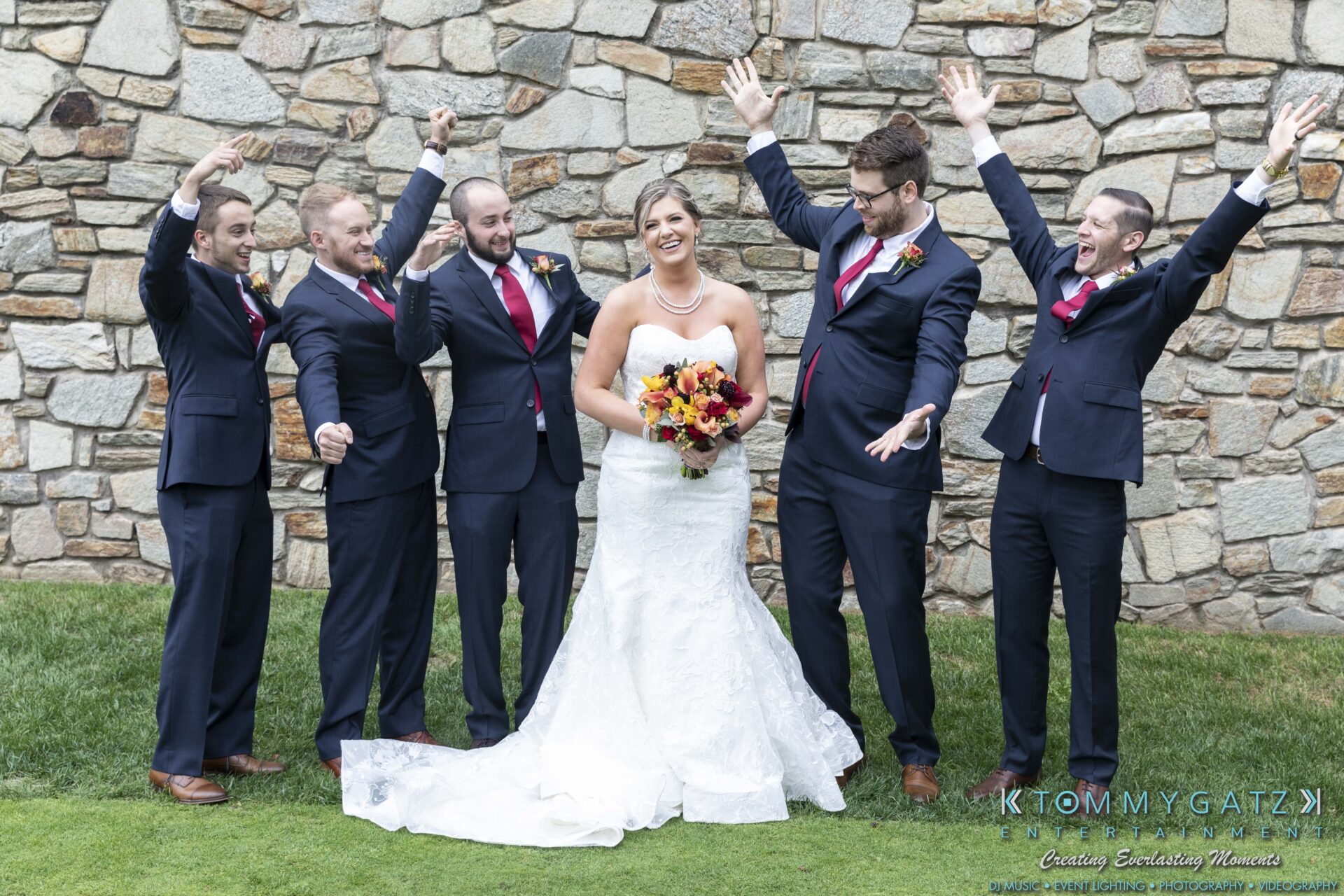 A bride in a white gown holding a bouquet stands with five groomsmen in navy suits and pink ties against a stone wall, all celebrating with raised arms and smiles.