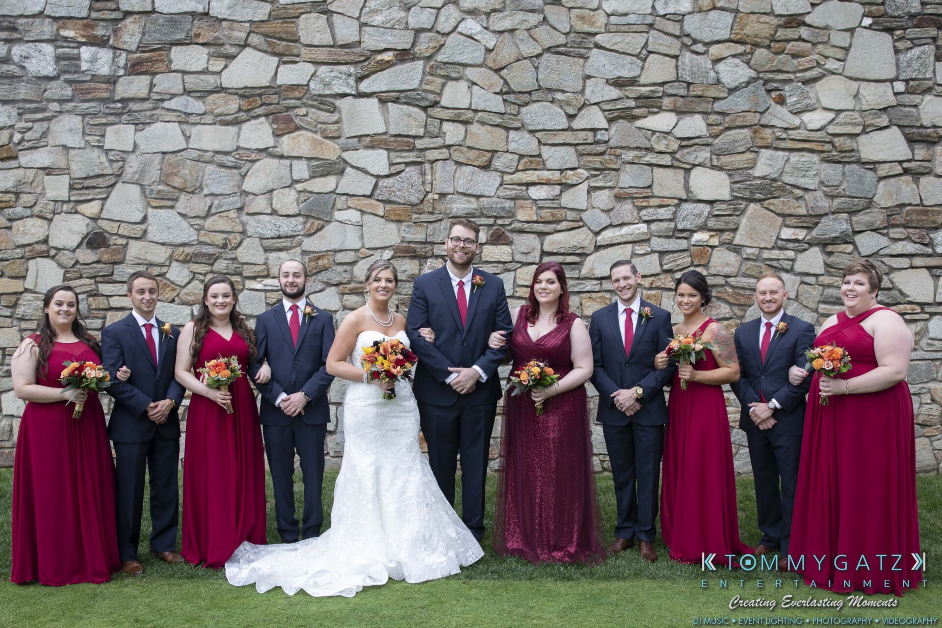 A wedding party with the bride and groom in the center, bridesmaids in red dresses, and groomsmen in dark suits, standing in front of a stone wall.