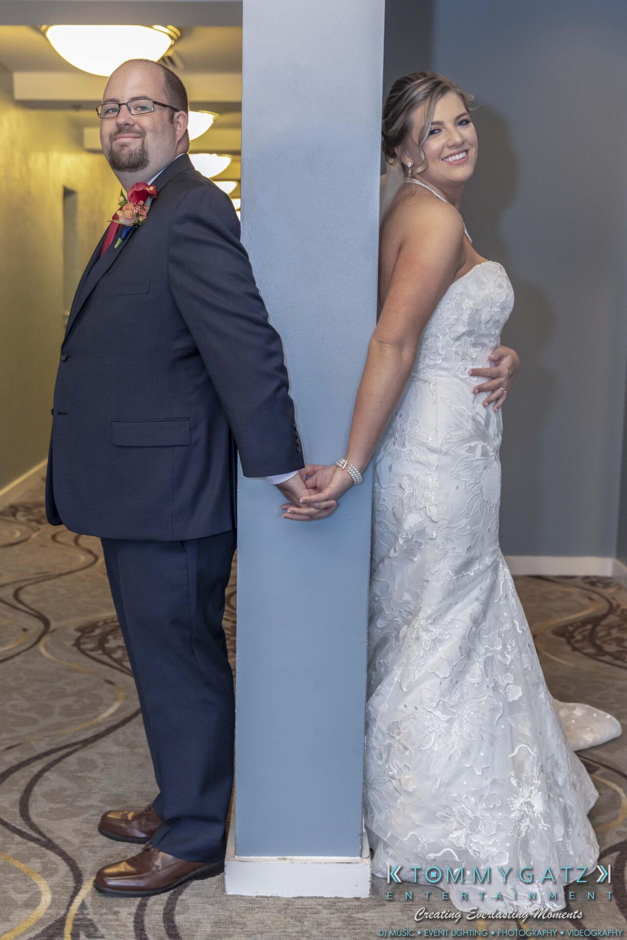 Bride and groom stand back-to-back, holding hands around a corner. Bride wears a white dress; groom is in a dark suit.