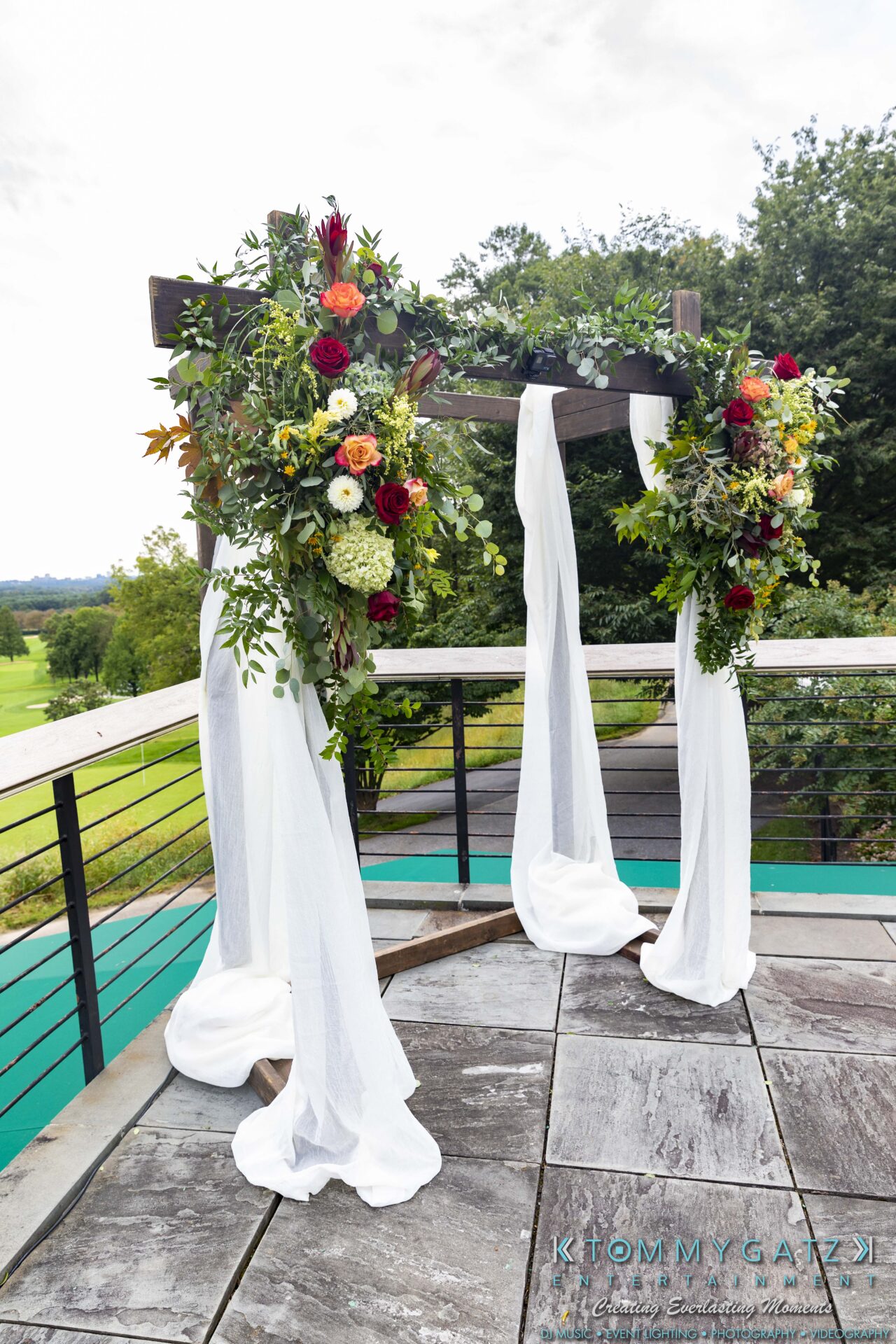 Wedding arch decorated with colorful flowers and white fabric, set on an outdoor deck overlooking greenery.