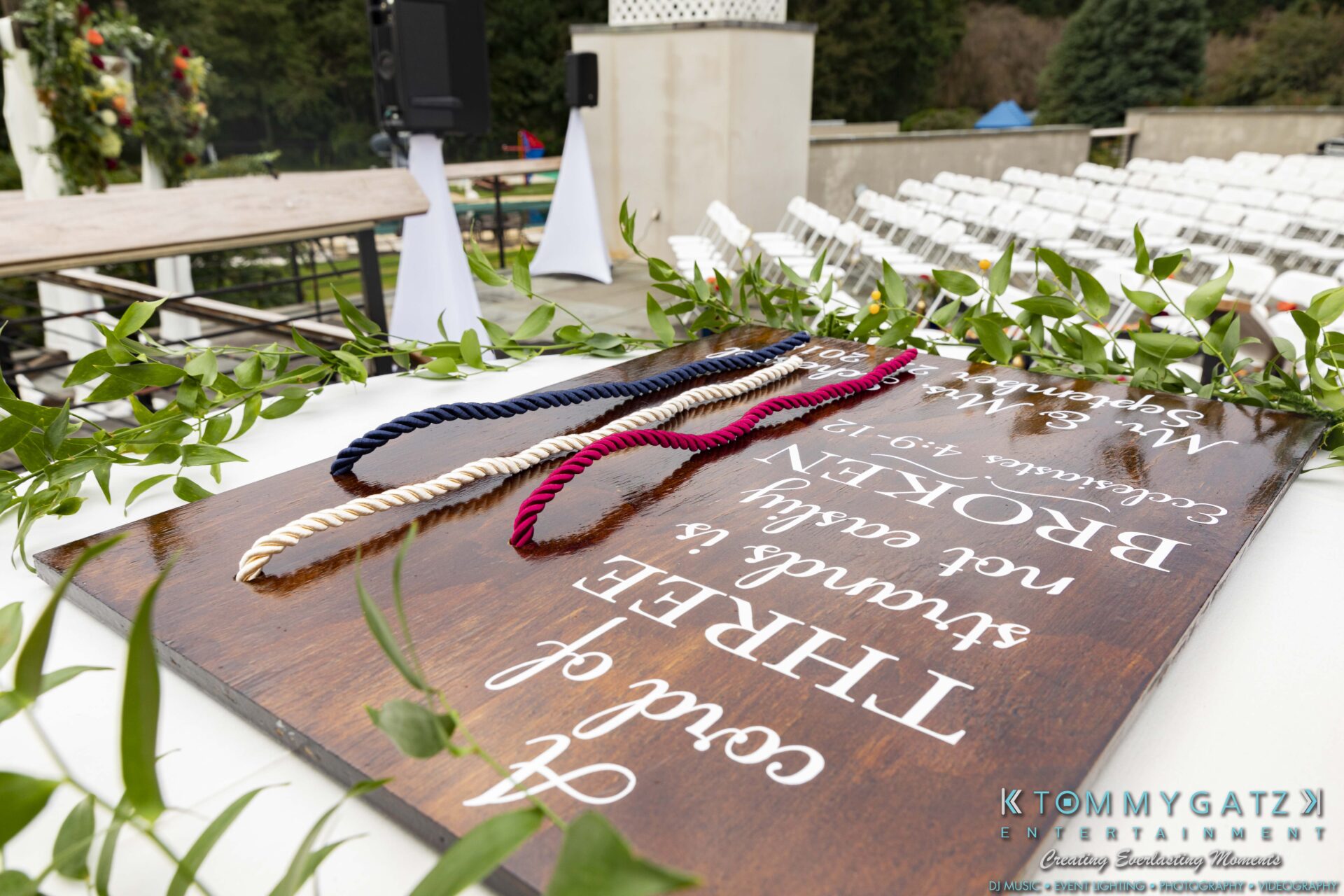 Wedding altar table with three colored cords on a wooden sign displaying a quote. White chairs and outdoor setting in the background.