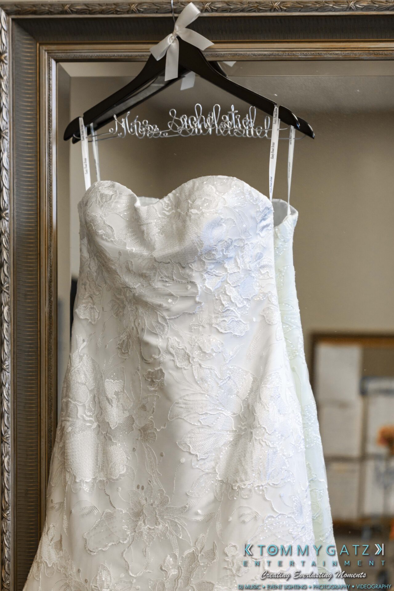 A white lace wedding dress hangs on a personalized hanger in front of a mirror with a decorative frame.