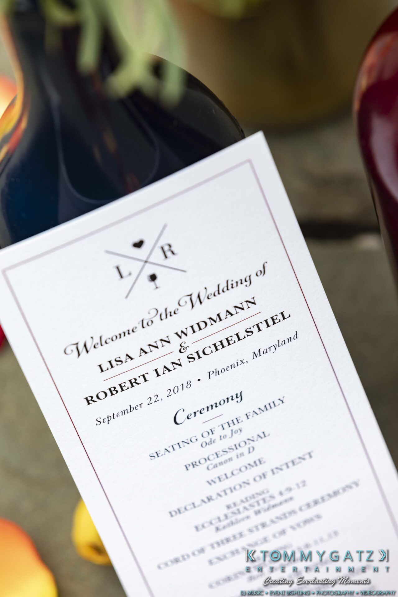 Close-up of a wedding program featuring the names Lisa Ann Widmann and Robert Ian Sichels­tiel, dated September 22, 2018, in Phoenix, Maryland, placed against a wine bottle.