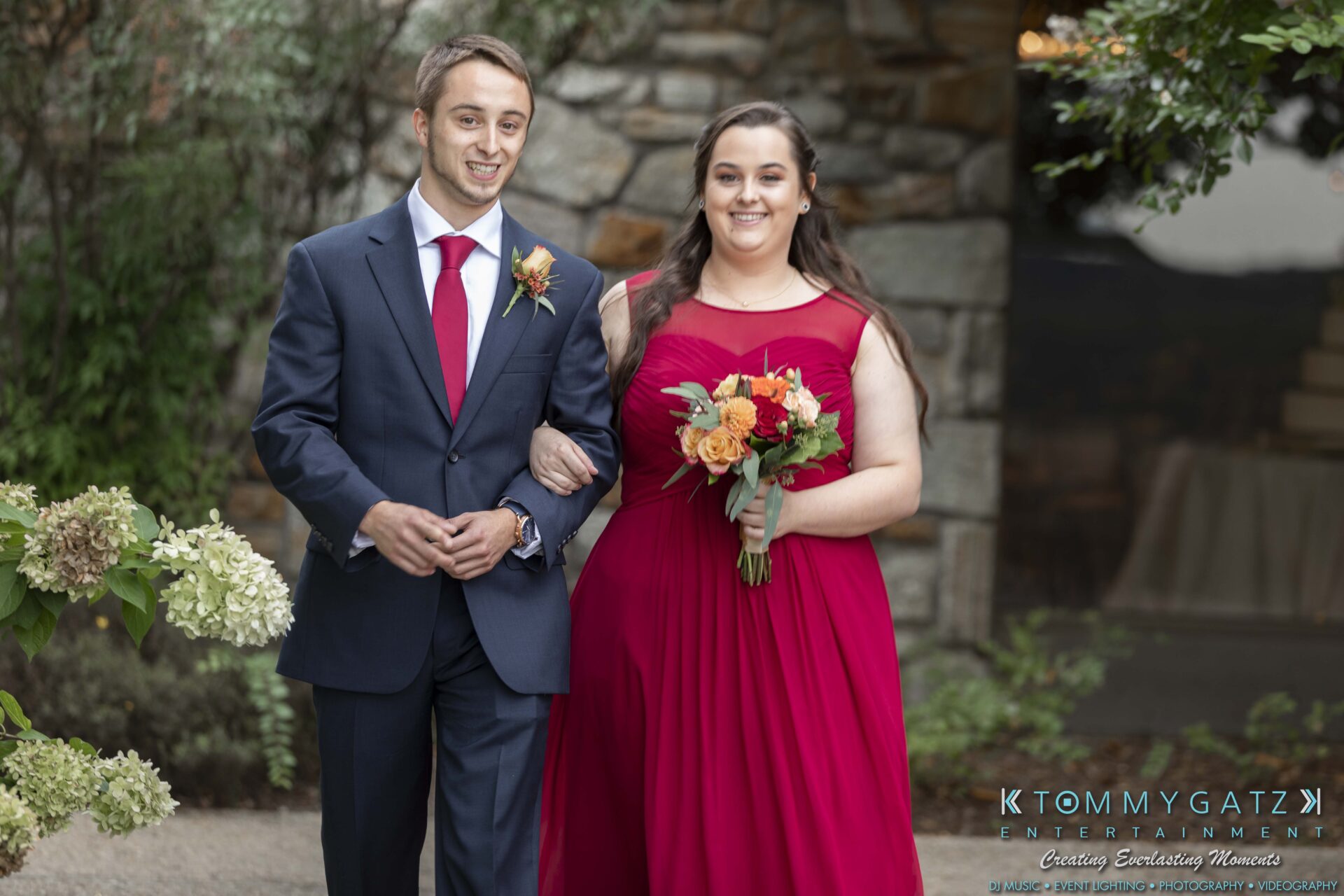 A man and woman in formal attire, with the woman holding a bouquet, walk arm in arm down a garden path.