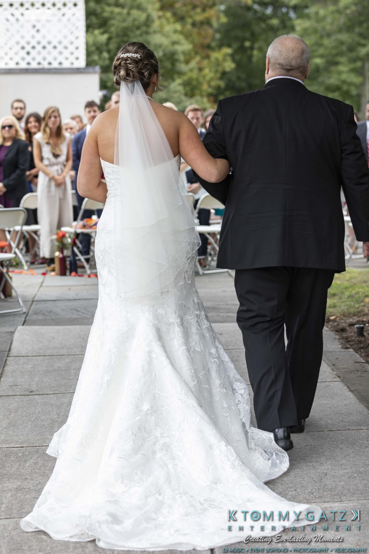 Bride in a white gown and veil walks down an outdoor aisle with an older man, while guests watch in the background.