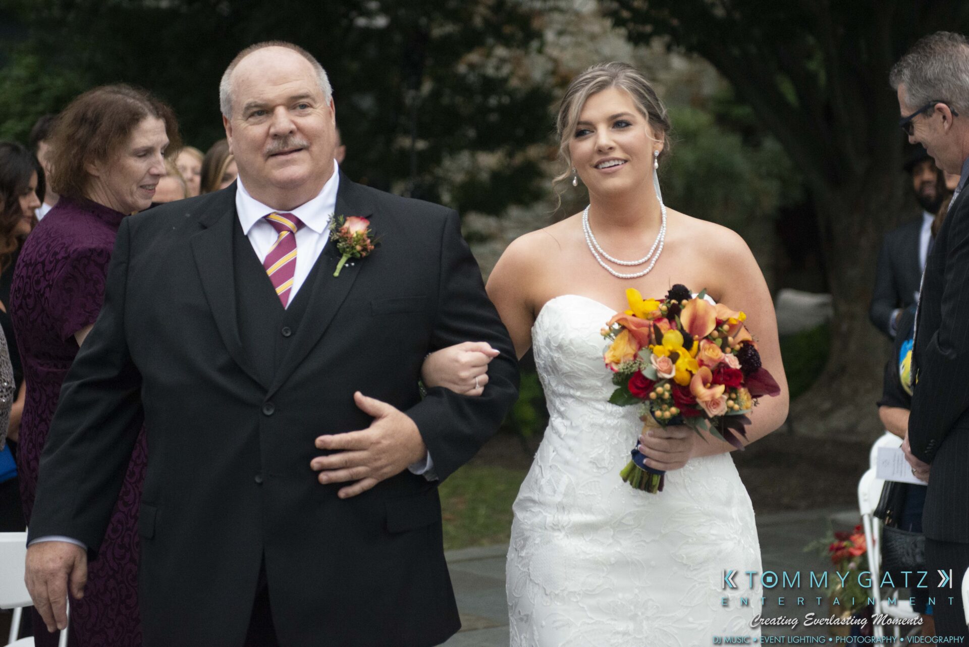 A bride in a white dress and pearl necklace walks down an outdoor aisle with an older man in a suit. She holds a bouquet of colorful flowers.