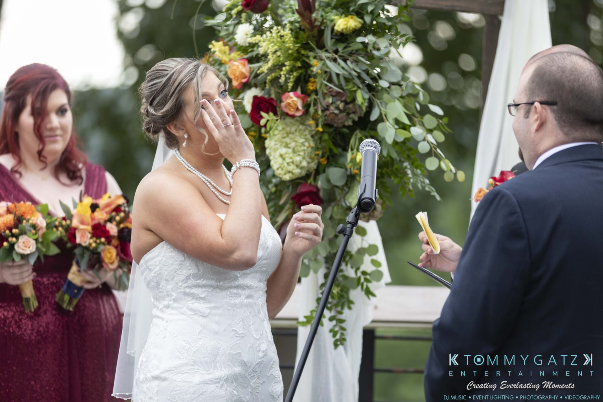 A bride wipes a tear during an outdoor wedding ceremony. A groom reads from notes. A bridesmaid holds a bouquet nearby.