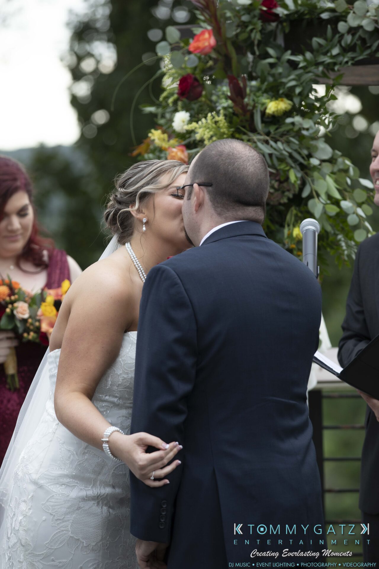 A bride and groom share a kiss during their wedding ceremony, with a partially visible officiant in the background holding flowers.
