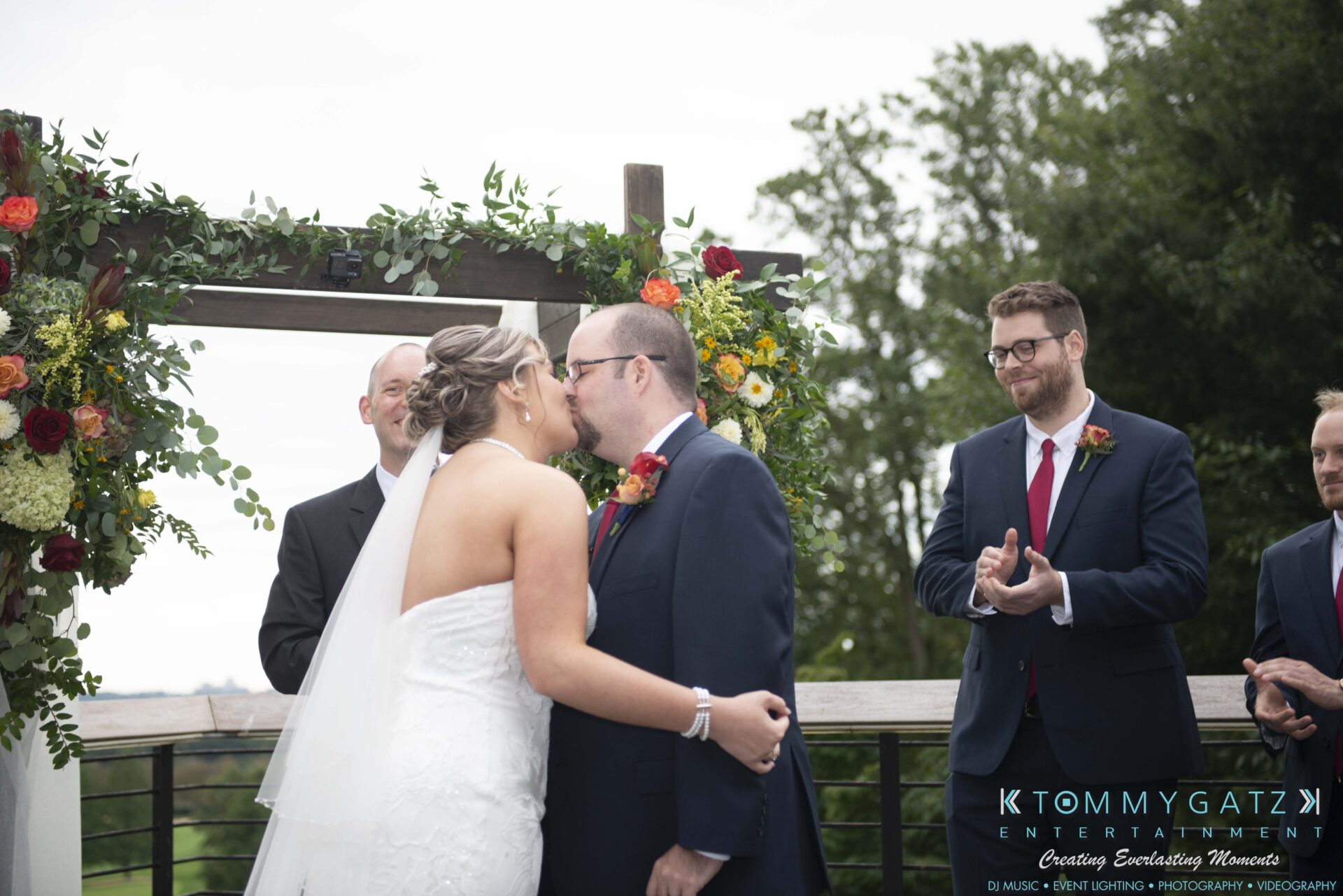 A bride and groom kiss at an outdoor wedding ceremony, with attendants clapping. Floral arch and greenery are visible.