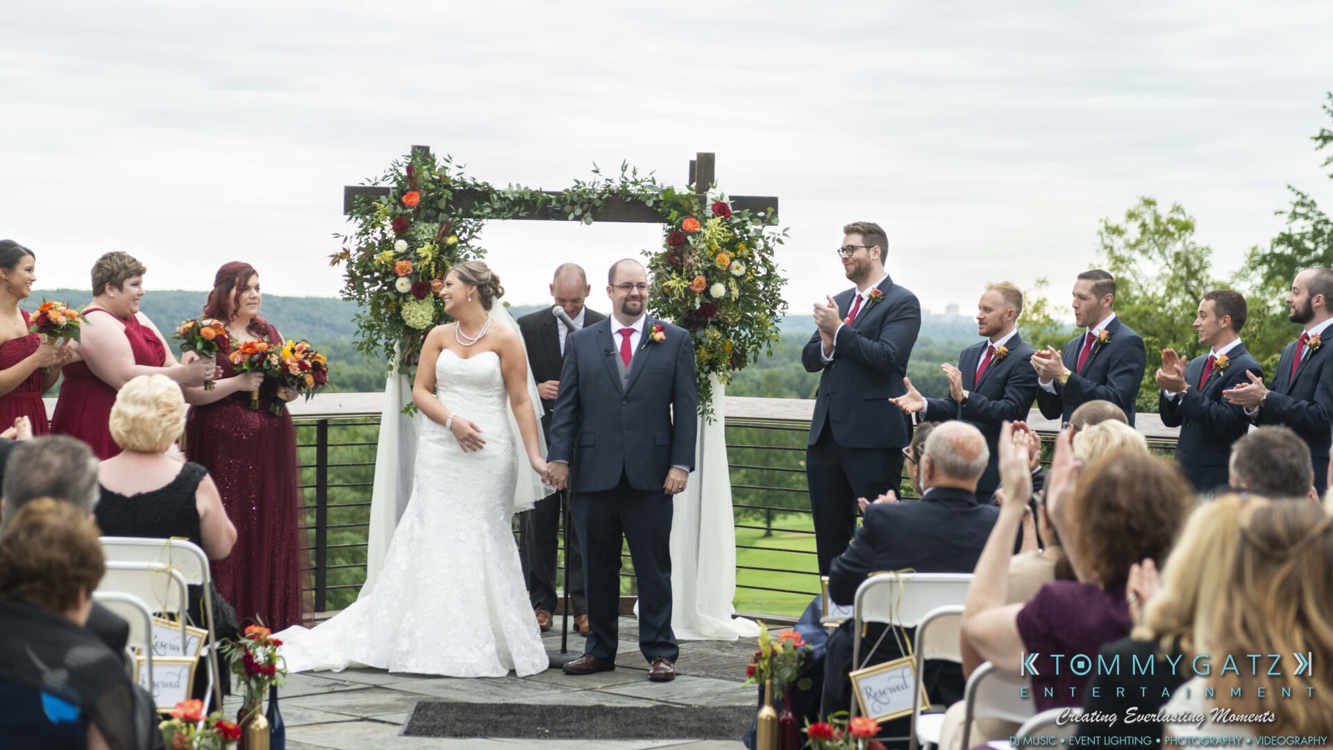 A bride and groom stand together at an outdoor wedding ceremony, surrounded by a bridal party and guests applauding. A floral arch and scenic landscape are in the background.