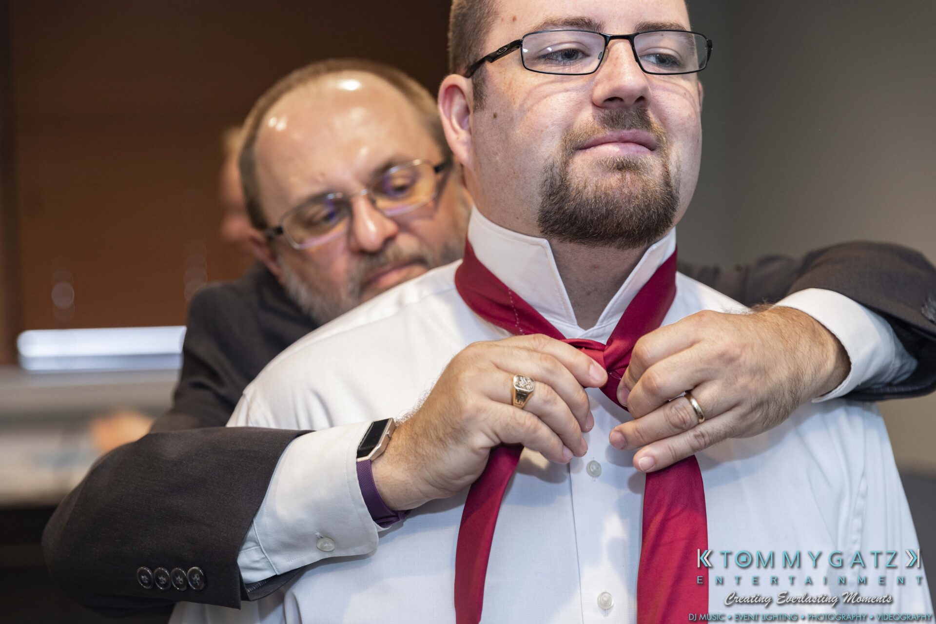 A man helps another man adjust a red bow tie, both wearing glasses and formal attire.