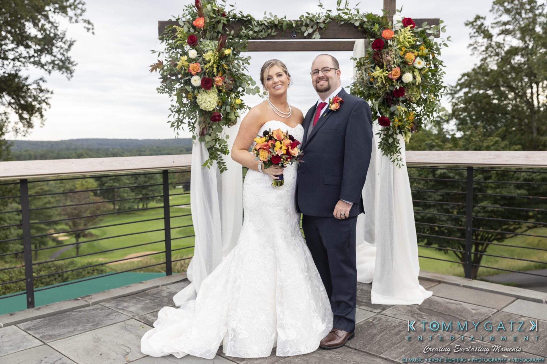 A couple poses in wedding attire under a floral arch on a terrace, with a scenic view in the background.