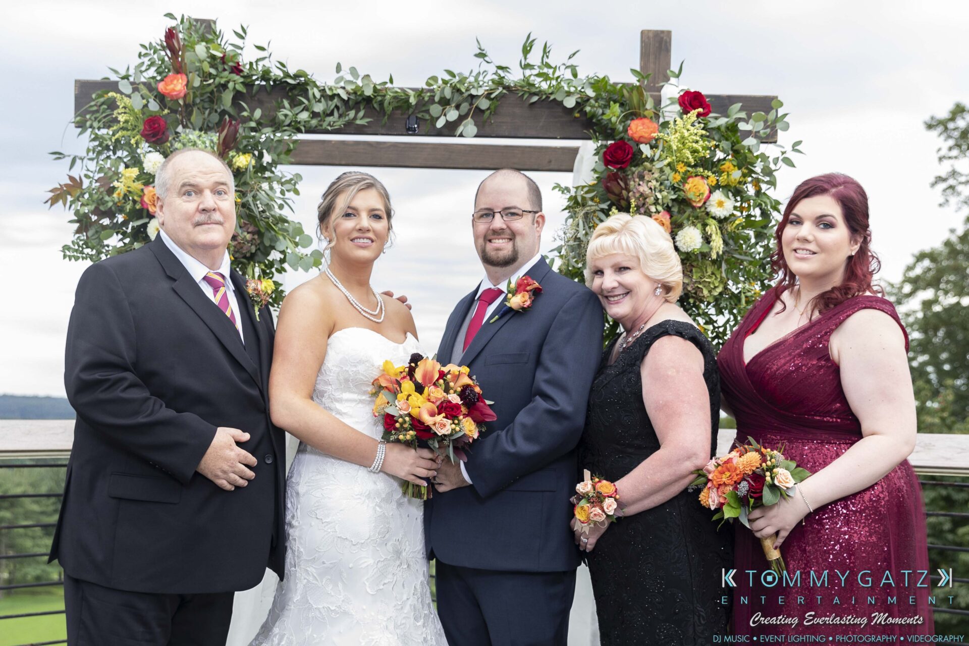 A wedding group photo with two couples and one woman in formal attire, standing in front of a floral archway outdoors.