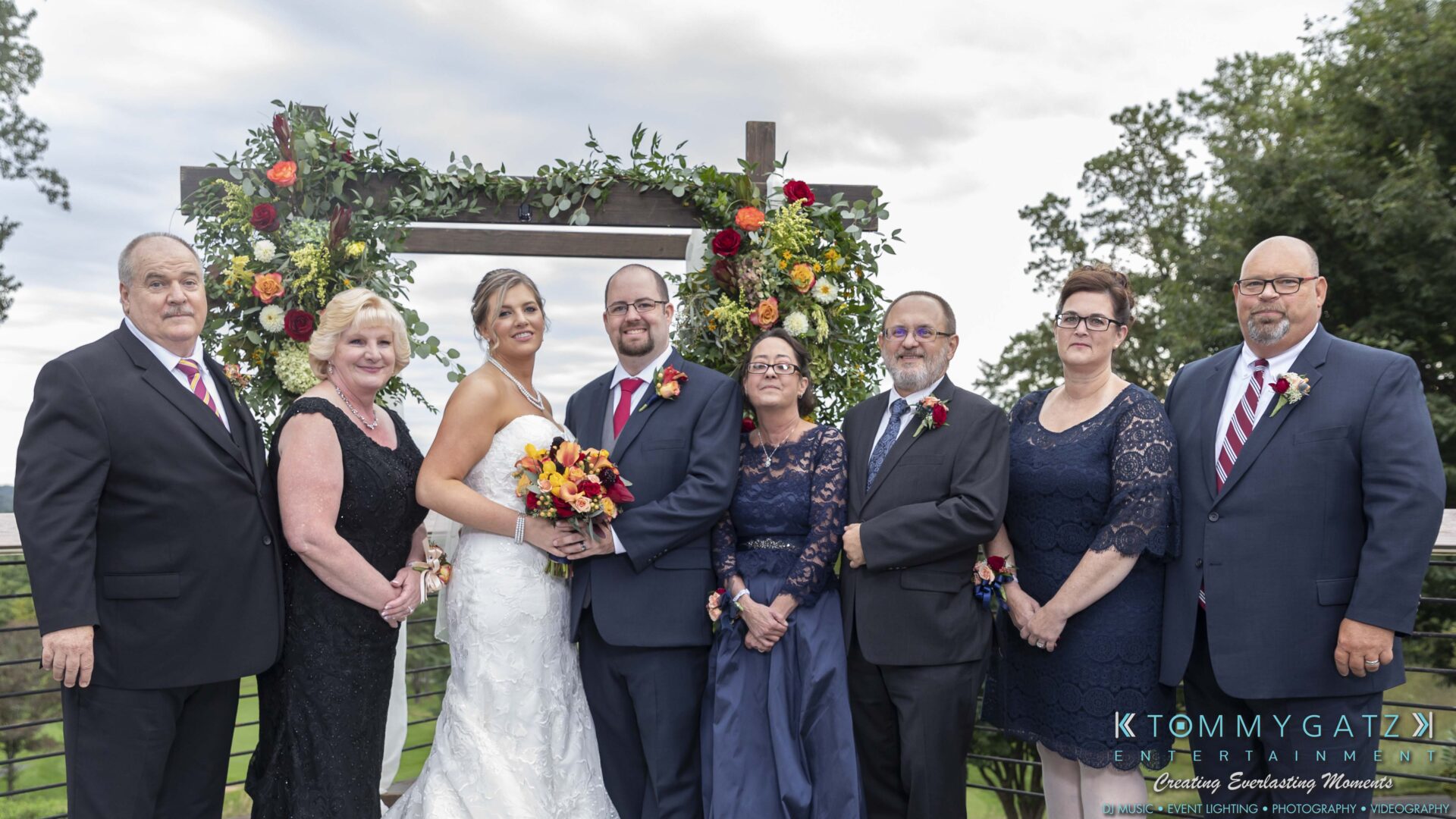 A wedding group photo with the bride, groom, and six relatives standing in front of a floral arch outdoors.