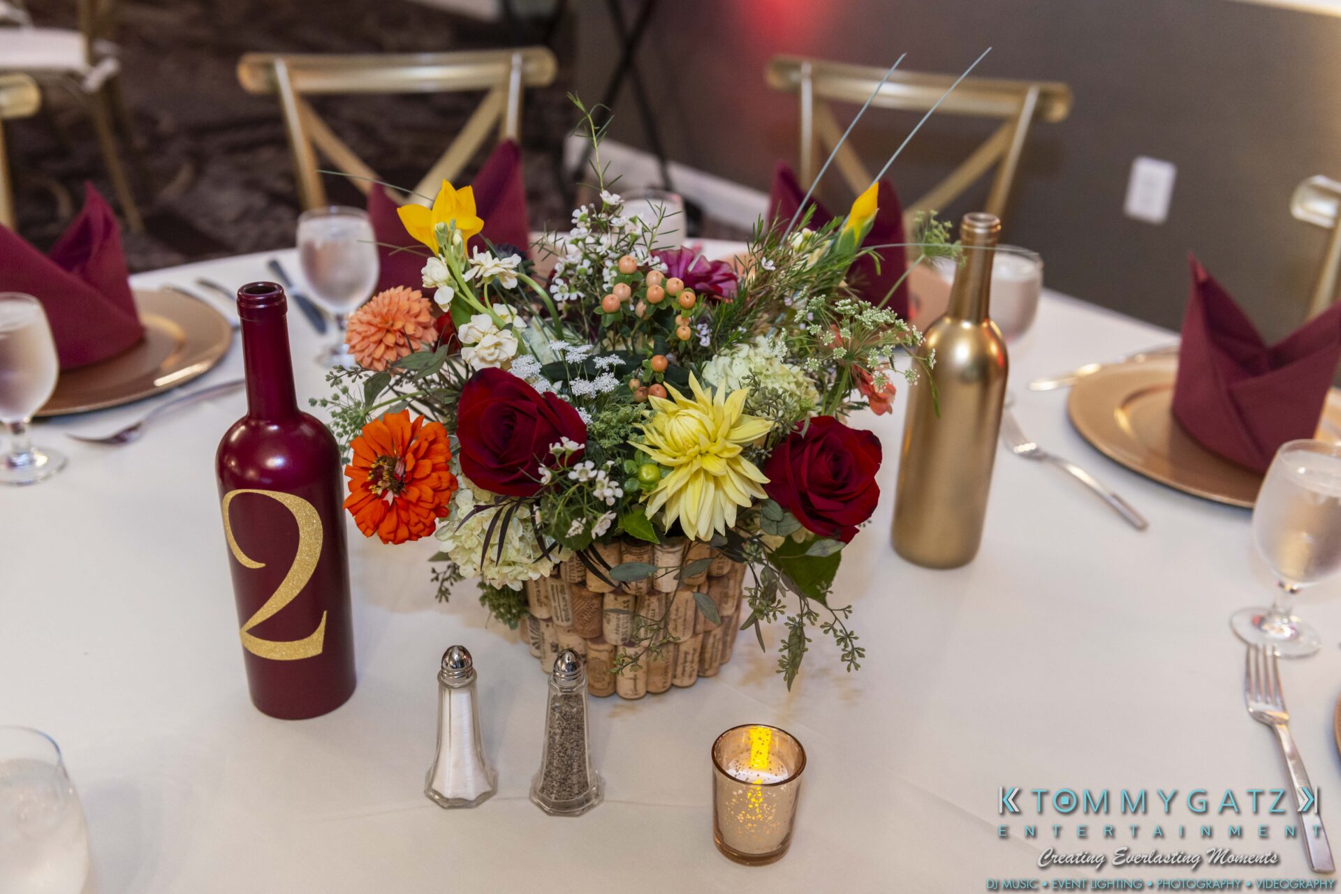 Table centerpiece with colorful flowers in a vase made of wine corks. Two wine bottles, one red with the number two, and gold one. Set dining table with folded napkins and utensils.