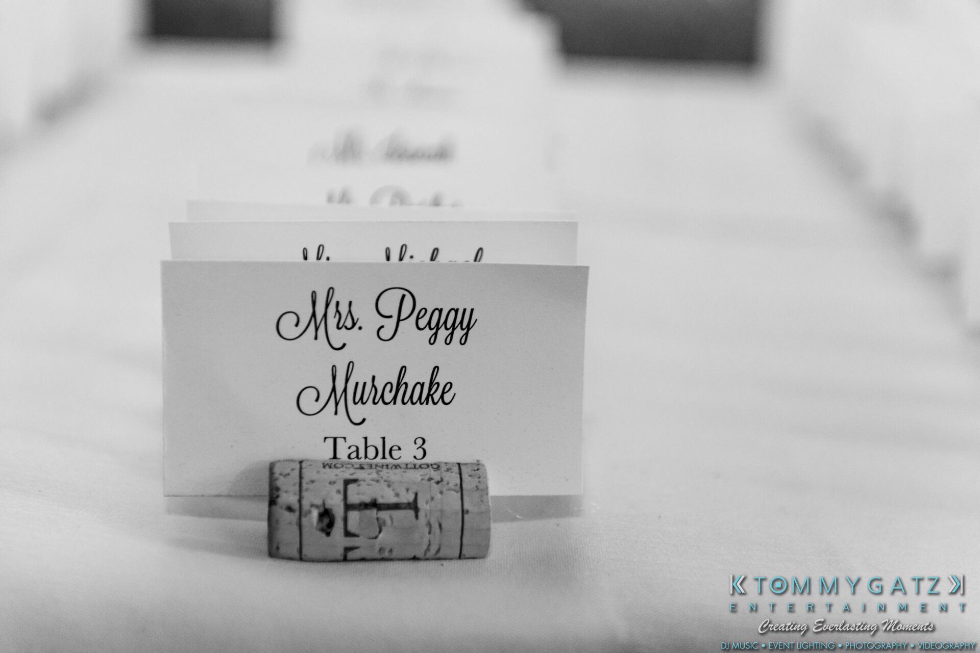 Place card on a table displays the name "Mrs. Peggy Murchake" and "Table 3," held by a wine cork. Tommy Gatz Entertainment watermark visible in the bottom right corner.