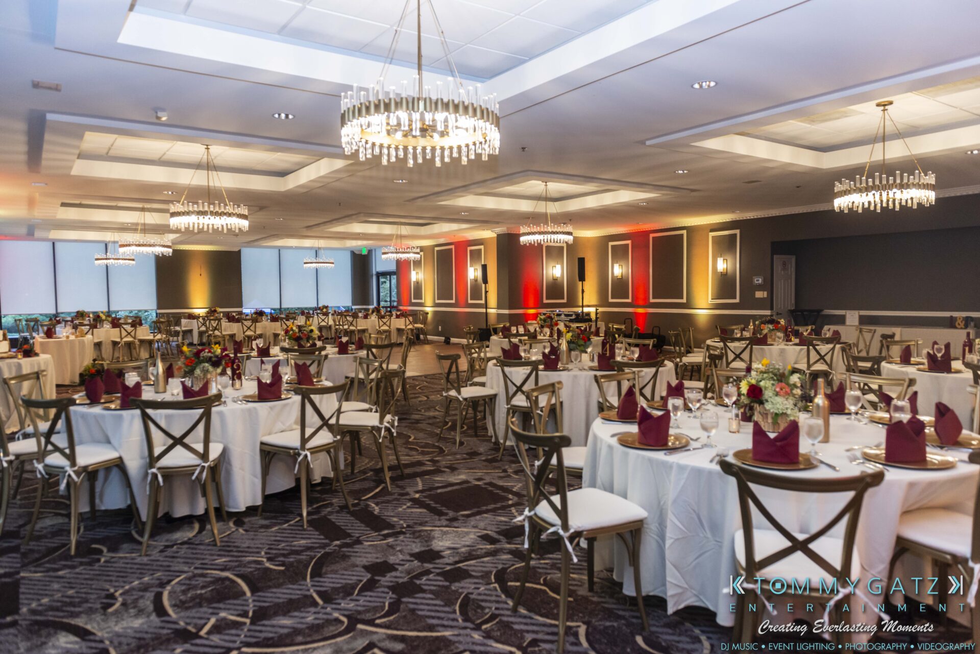 A banquet hall with round tables set for an event, featuring white tablecloths, burgundy napkins, and wooden chairs. Chandeliers hang from the ceiling, providing elegant lighting.