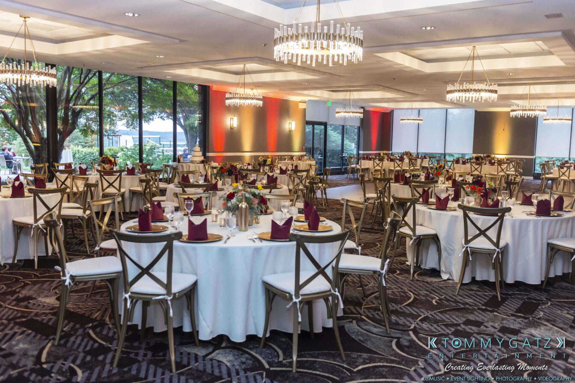 Elegant banquet hall with round tables set for an event, featuring white tablecloths, burgundy napkins, and floral centerpieces, illuminated by chandeliers and natural light from large windows.