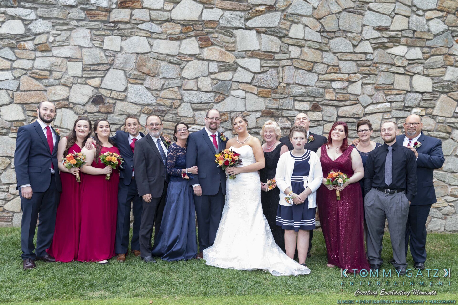 A group of people, including a bride and groom, pose in formal attire outdoors in front of a stone wall. Some hold bouquets.