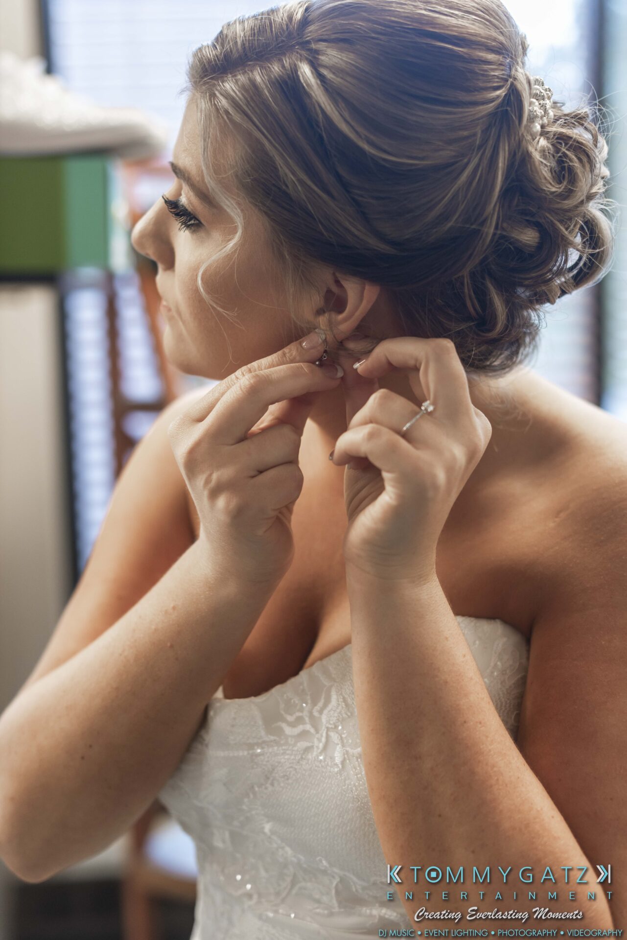 A bride in a white strapless dress adjusts her earring in front of a window.