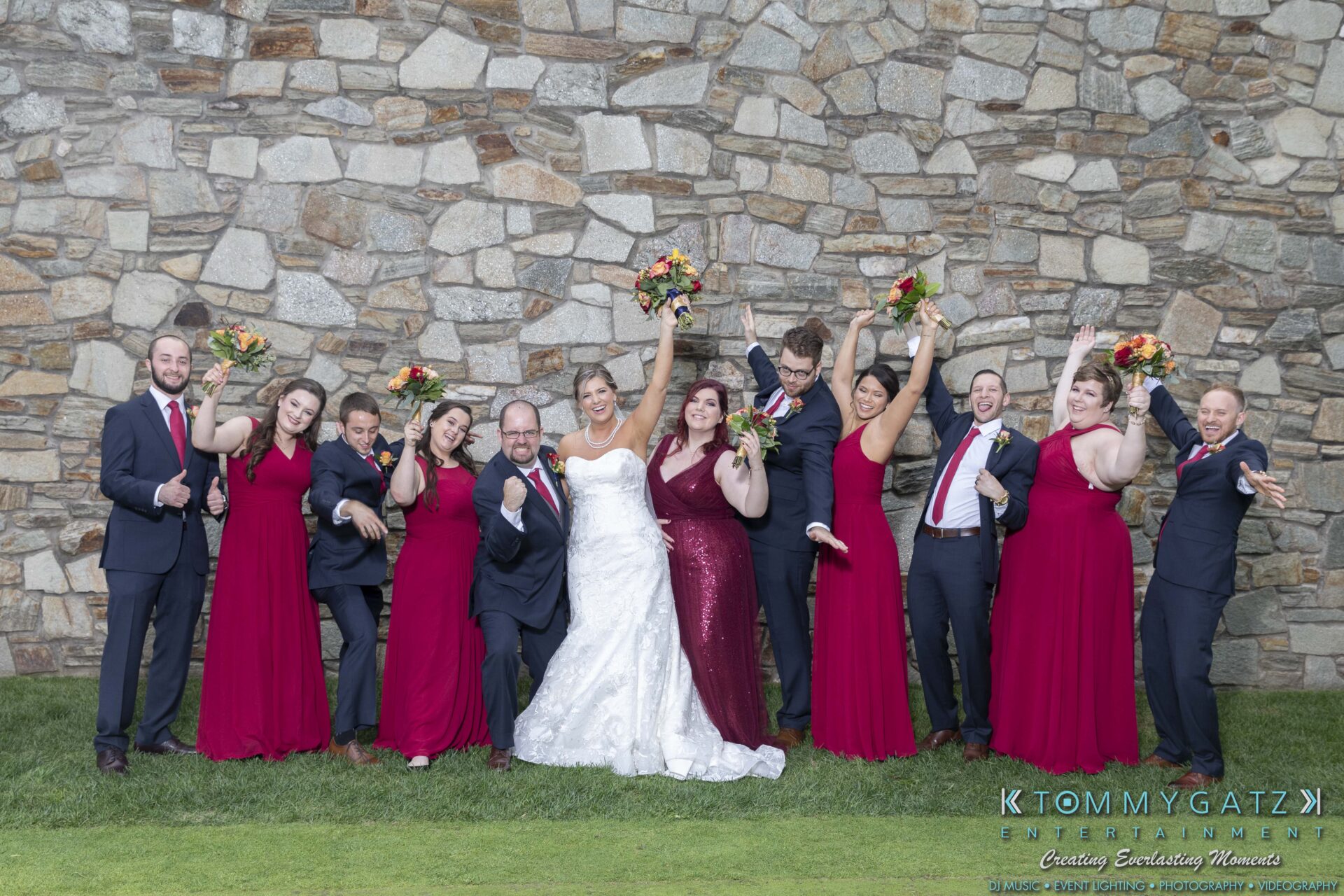 A wedding party poses outdoors against a stone wall. The bride and groom are in the center, surrounded by bridesmaids in burgundy dresses and groomsmen in black suits holding bouquets.