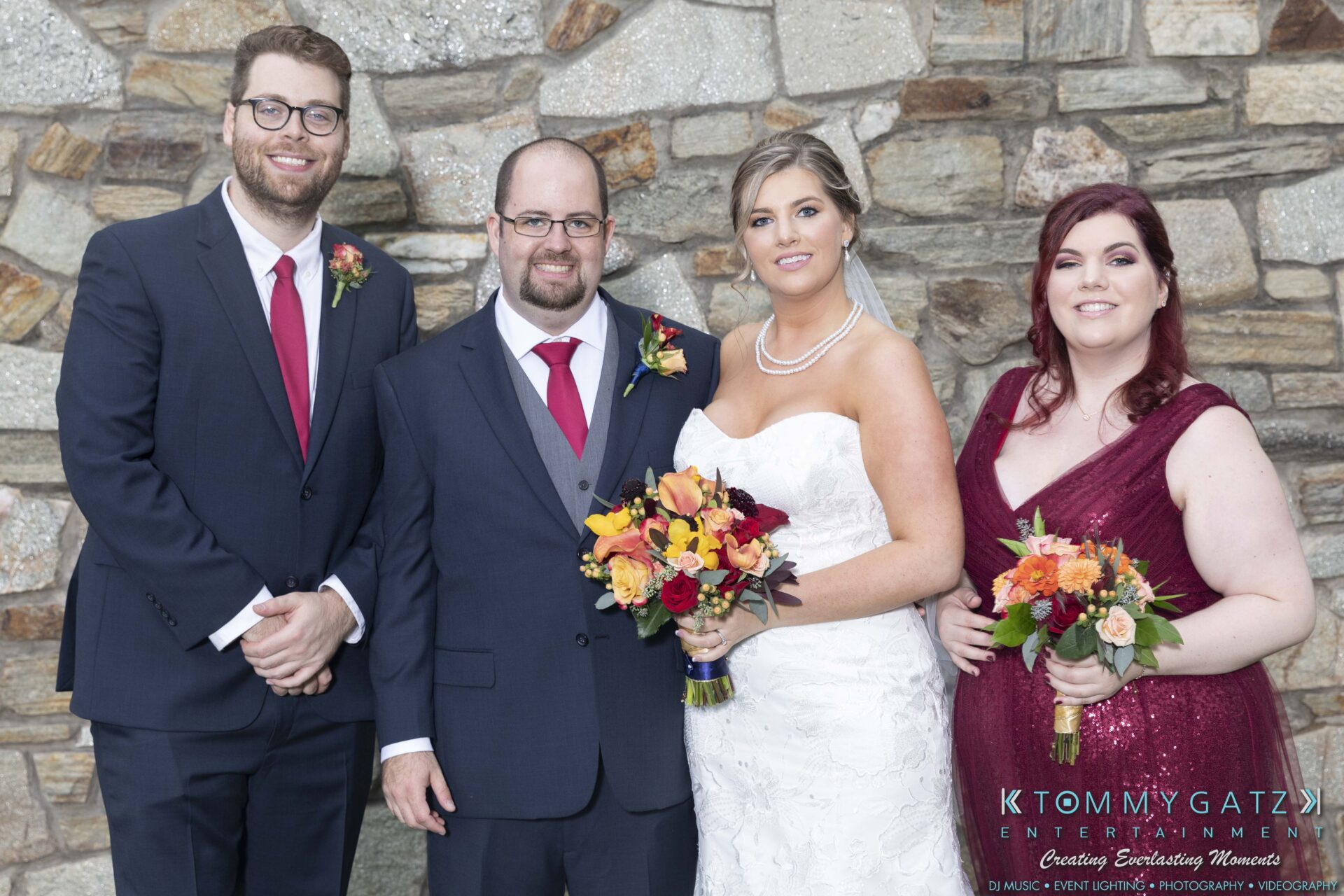 Four people pose in formal attire at an event, with two holding flower bouquets. A stone wall serves as the backdrop.