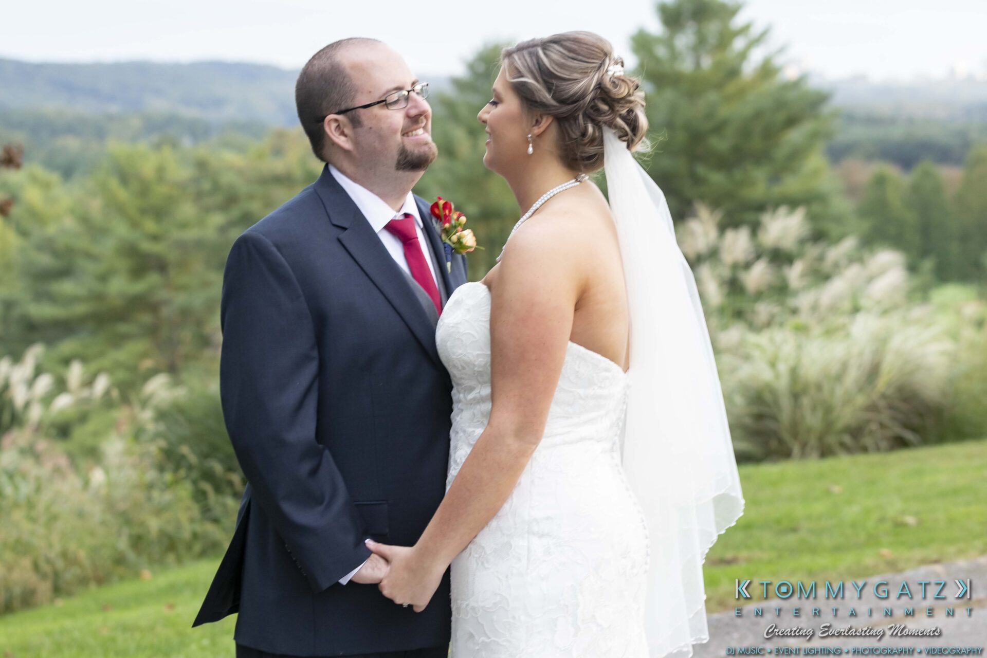 A bride and groom stand outdoors, holding hands and smiling at each other, with a scenic background of greenery and distant hills.