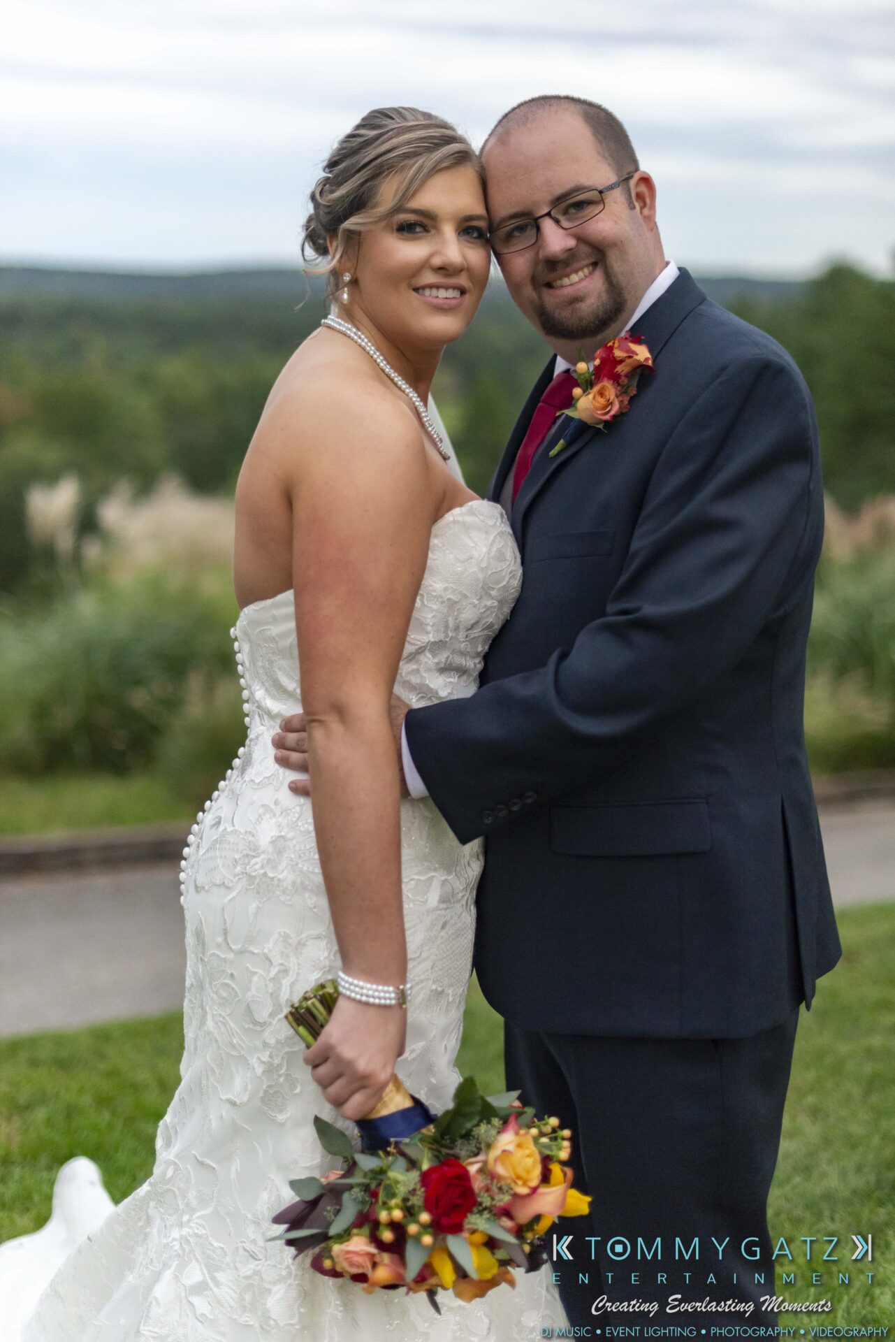 A couple poses outdoors on their wedding day. The woman is in a white gown holding a bouquet, and the man is in a dark suit with a boutonniere.