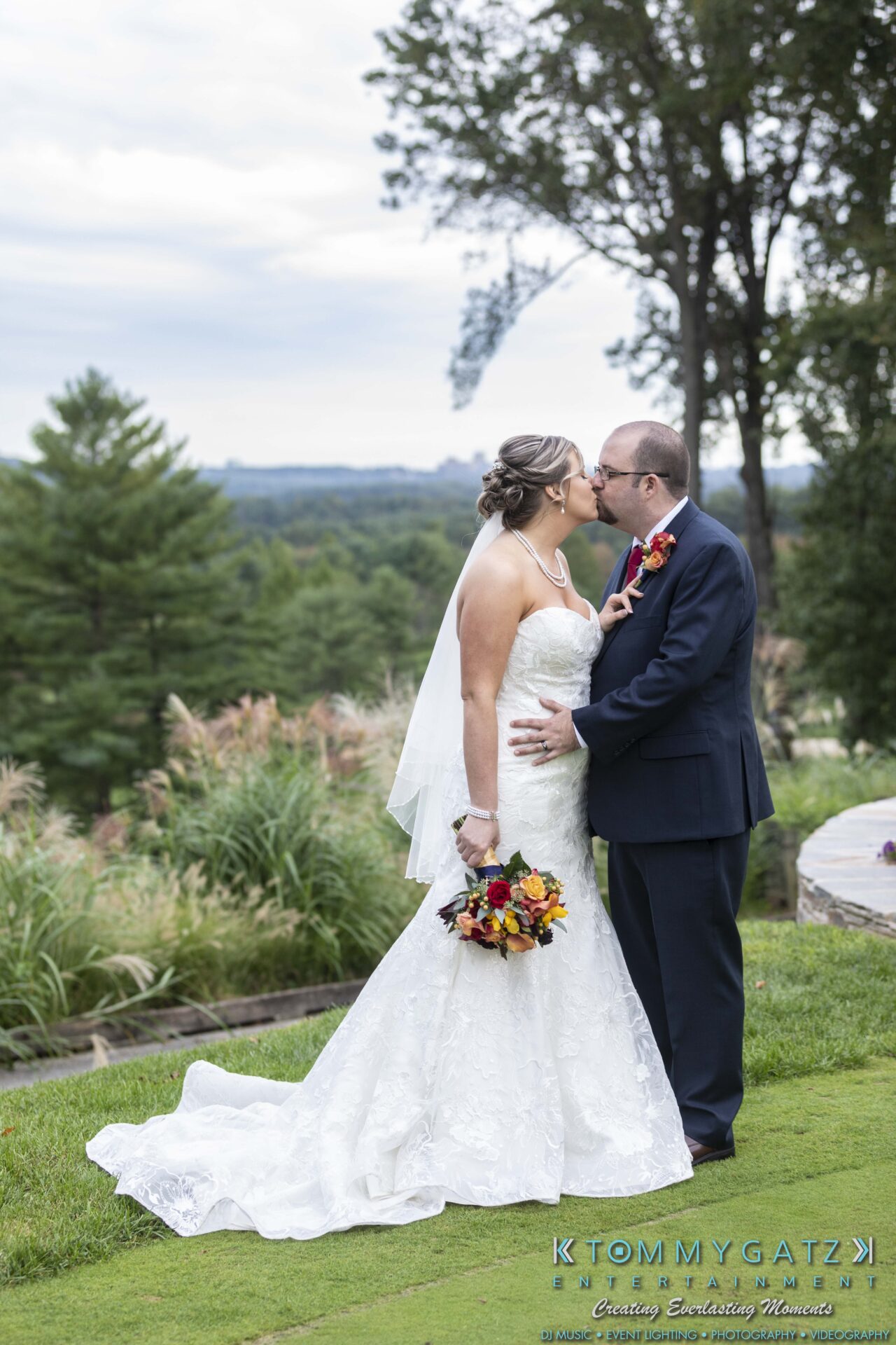 A bride and groom kissing outdoors in a garden setting. The bride holds a bouquet of colorful flowers, and the groom is in a dark suit. Trees and landscape are visible in the background.
