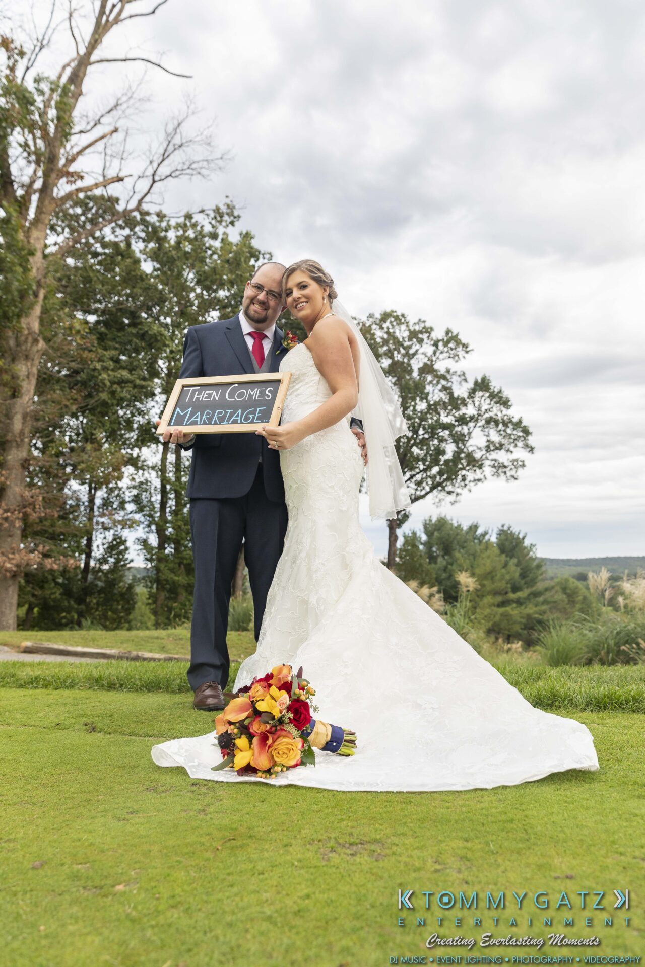 Bride and groom stand outdoors holding a sign reading, "Then comes marriage," with a bouquet on the grass in front of them.