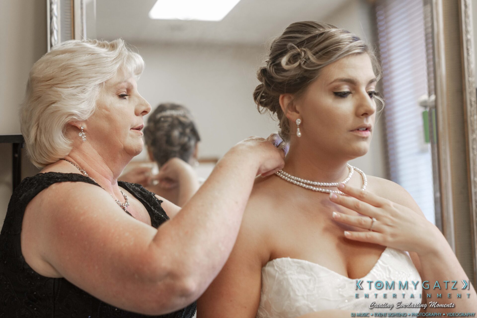 A woman helps another woman in a white dress adjust her necklace in front of a mirror.
