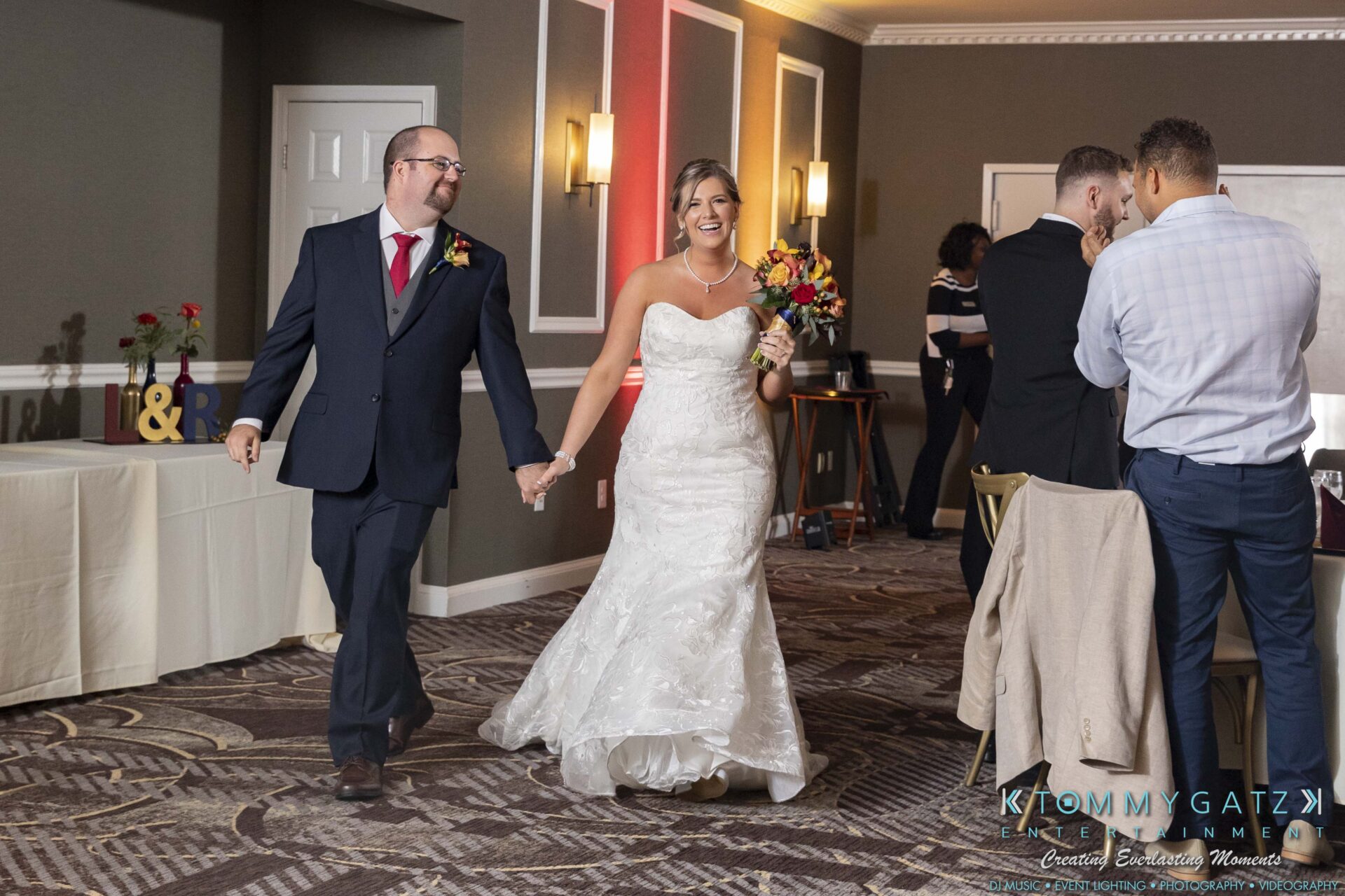 A bride and groom enter a decorated room, smiling and holding hands. The bride wears a white gown, and the groom is in a suit. Guests are visible in the background.