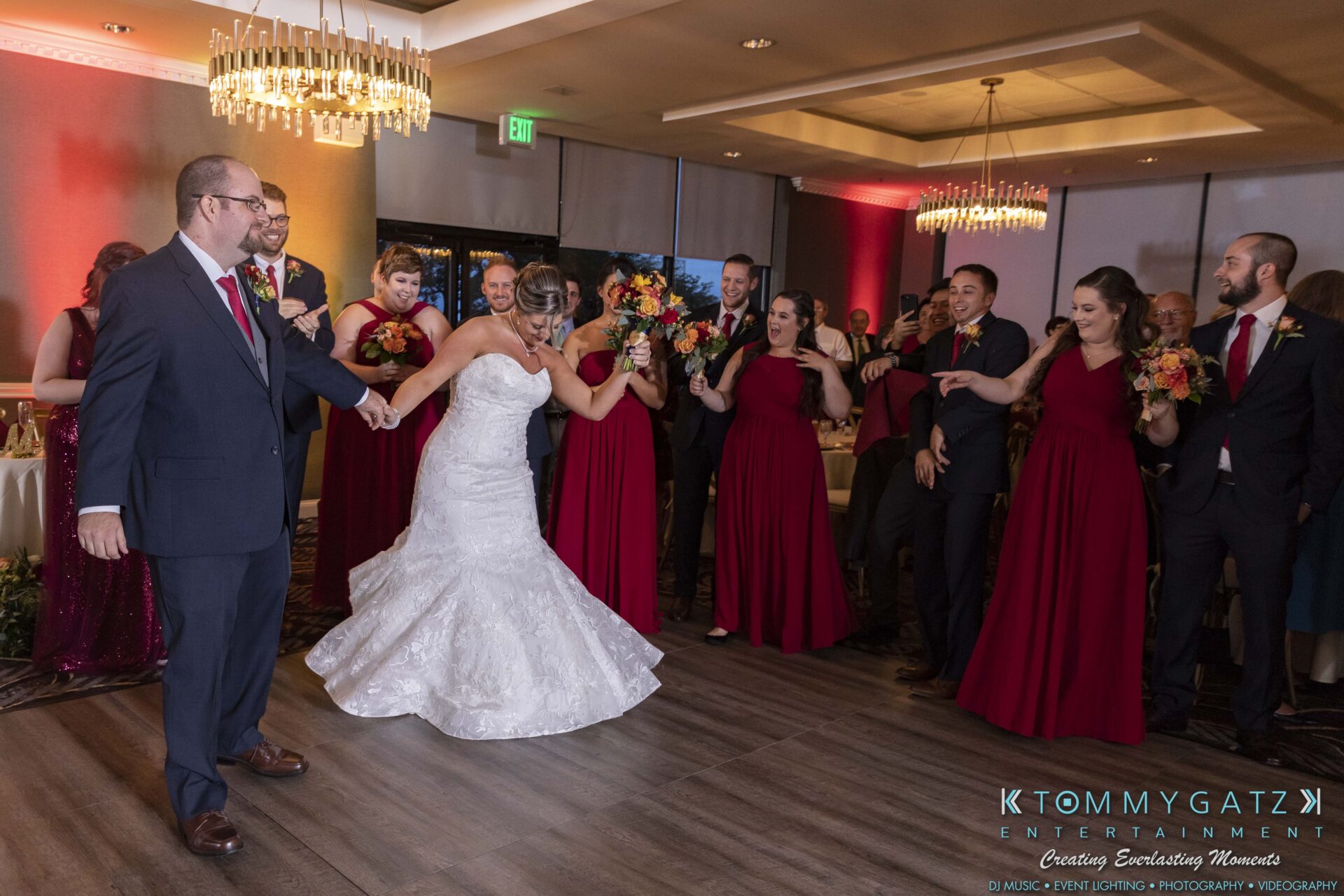 A bride in a white gown dances with a groom in a suit, surrounded by a group of people in red dresses and suits, inside a decorated event venue.