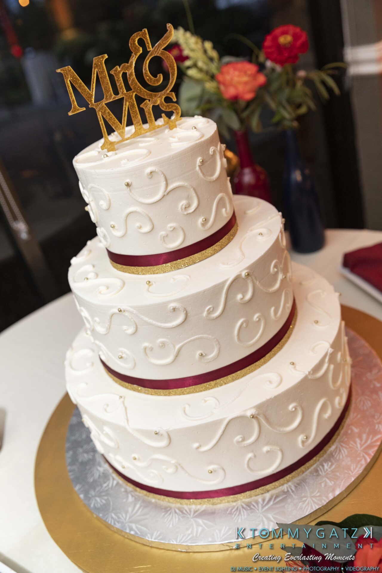 Three-tier white wedding cake with red ribbon accents and a "Mr & Mrs" topper, on a silver and gold base.