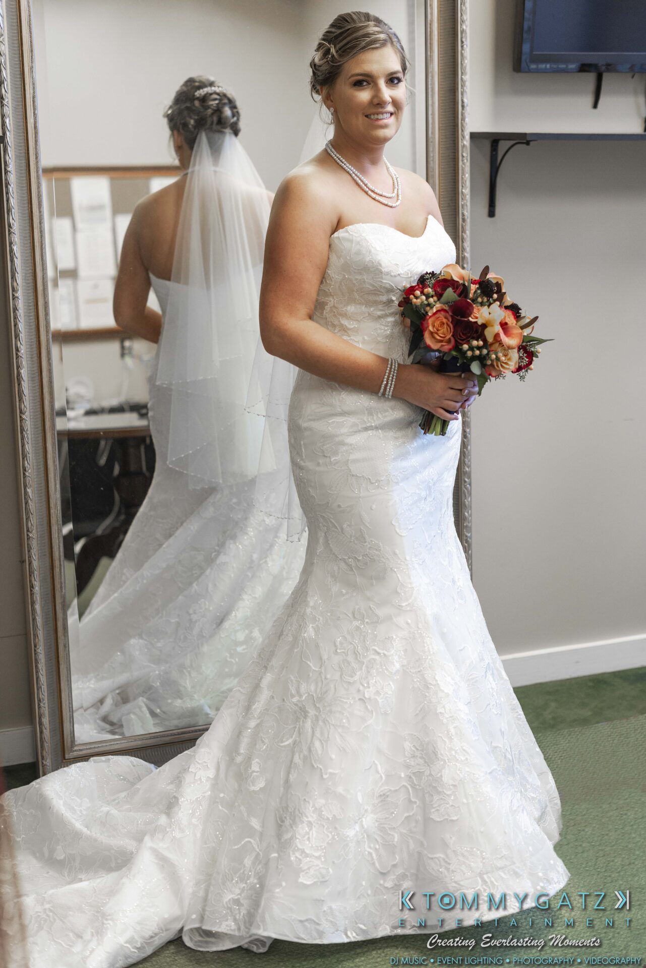 A bride in a white strapless wedding dress holds a bouquet and stands in front of a mirror, with her reflection visible.