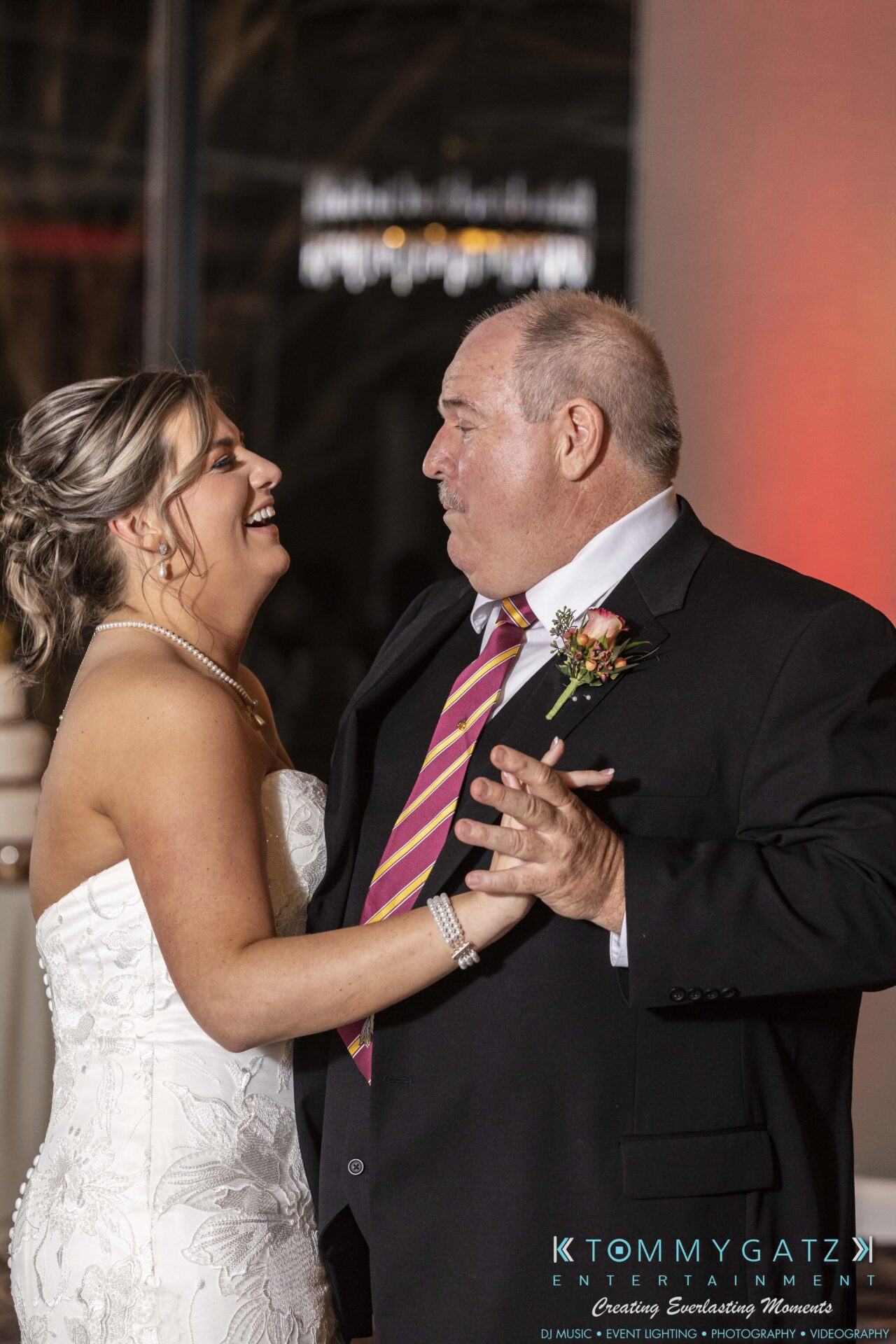 A bride in a white dress dances with an older man in a suit and striped tie at an indoor event.