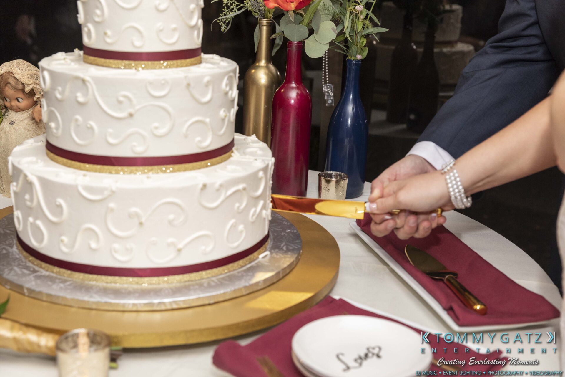 A couple cuts a three-tiered white cake with a gold knife. The table is set with plates and red napkins.