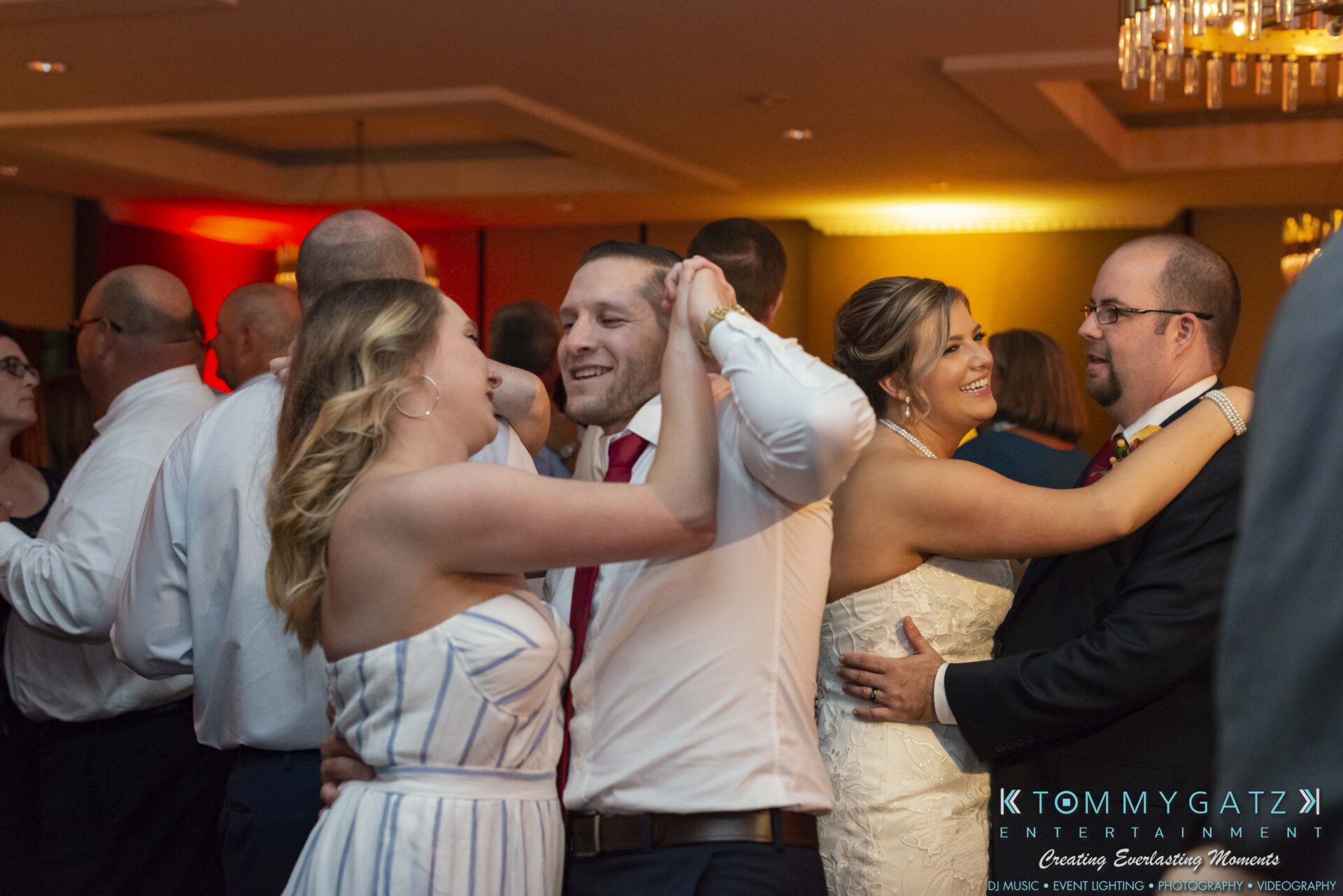 People dancing in pairs at an indoor social gathering. The room is warmly lit, and everyone appears to be enjoying the event.