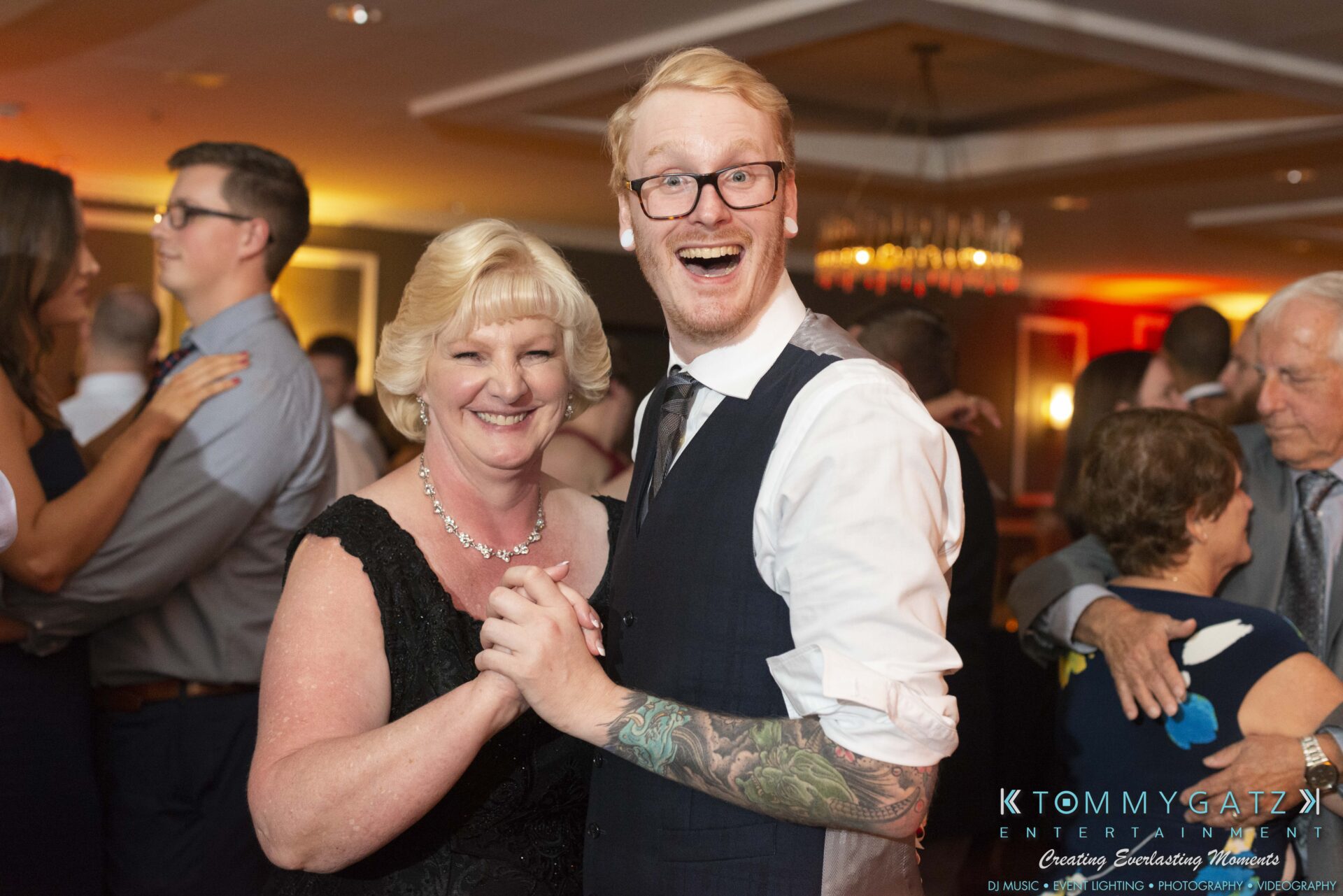 A man and a woman smiling while dancing at an event, with other attendees in the background.