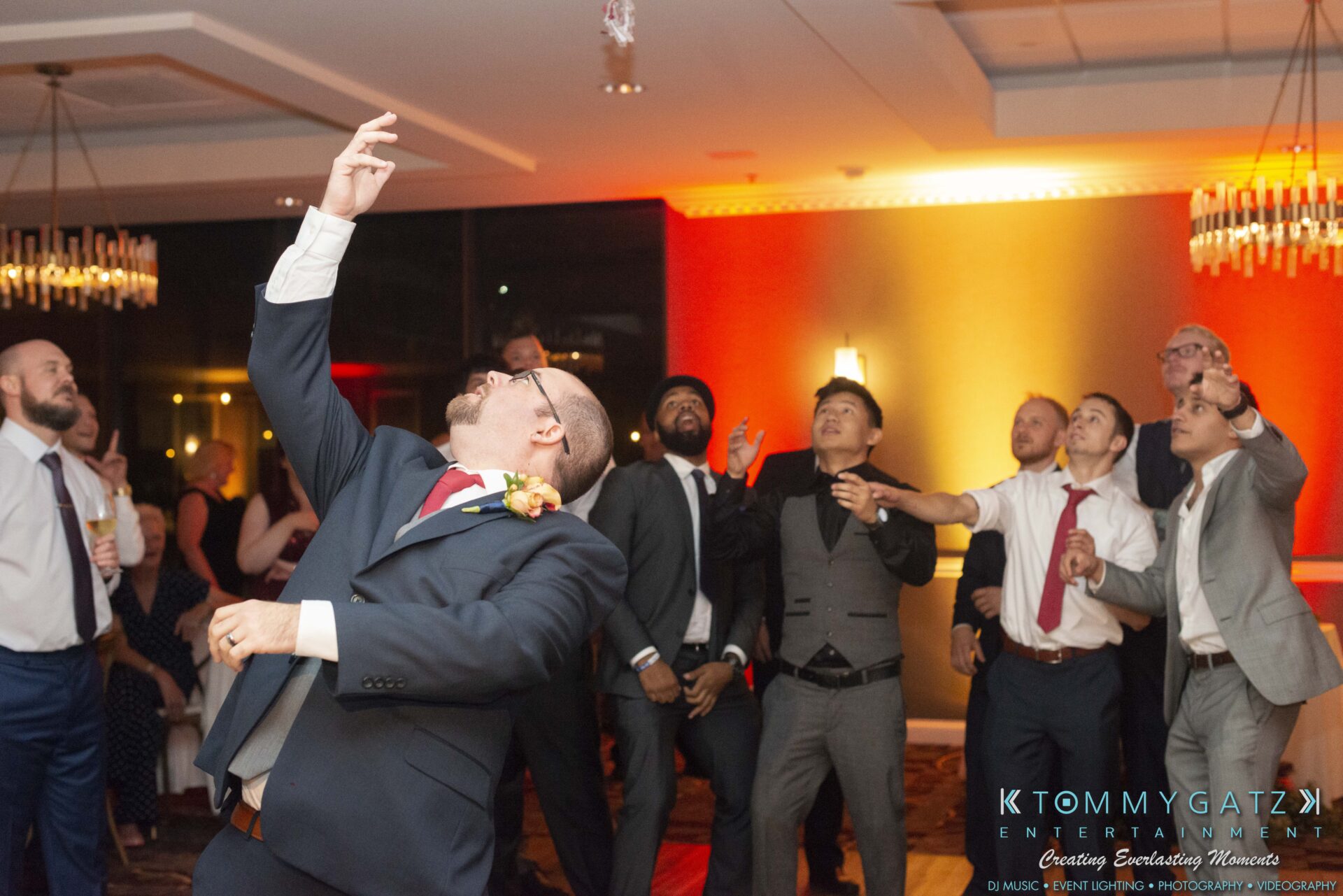 A group of people in formal attire are enthusiastically reaching upwards at an indoor event with colorful lighting.