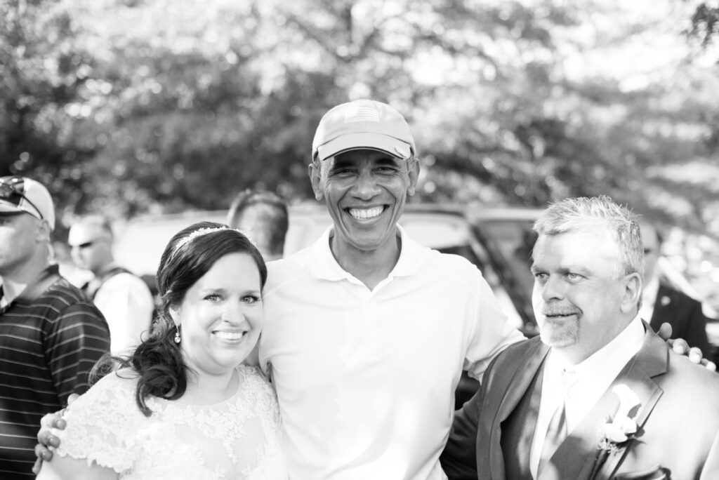 A person in a cap stands smiling between a bride in a lace dress and a man in a suit outdoors.